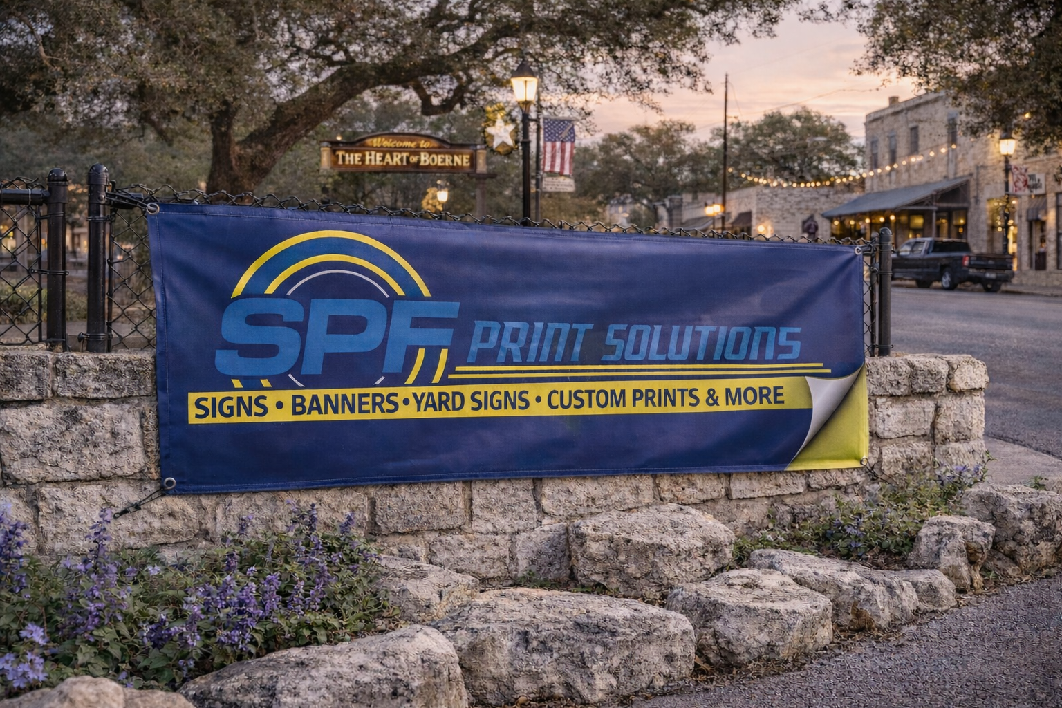 A blue banner advertising SPF Print Solutions with yellow and blue text, displayed outdoors on a stone wall in a small town, with a sign reading "The Heart of Boerne" and historic buildings in the background at dusk.