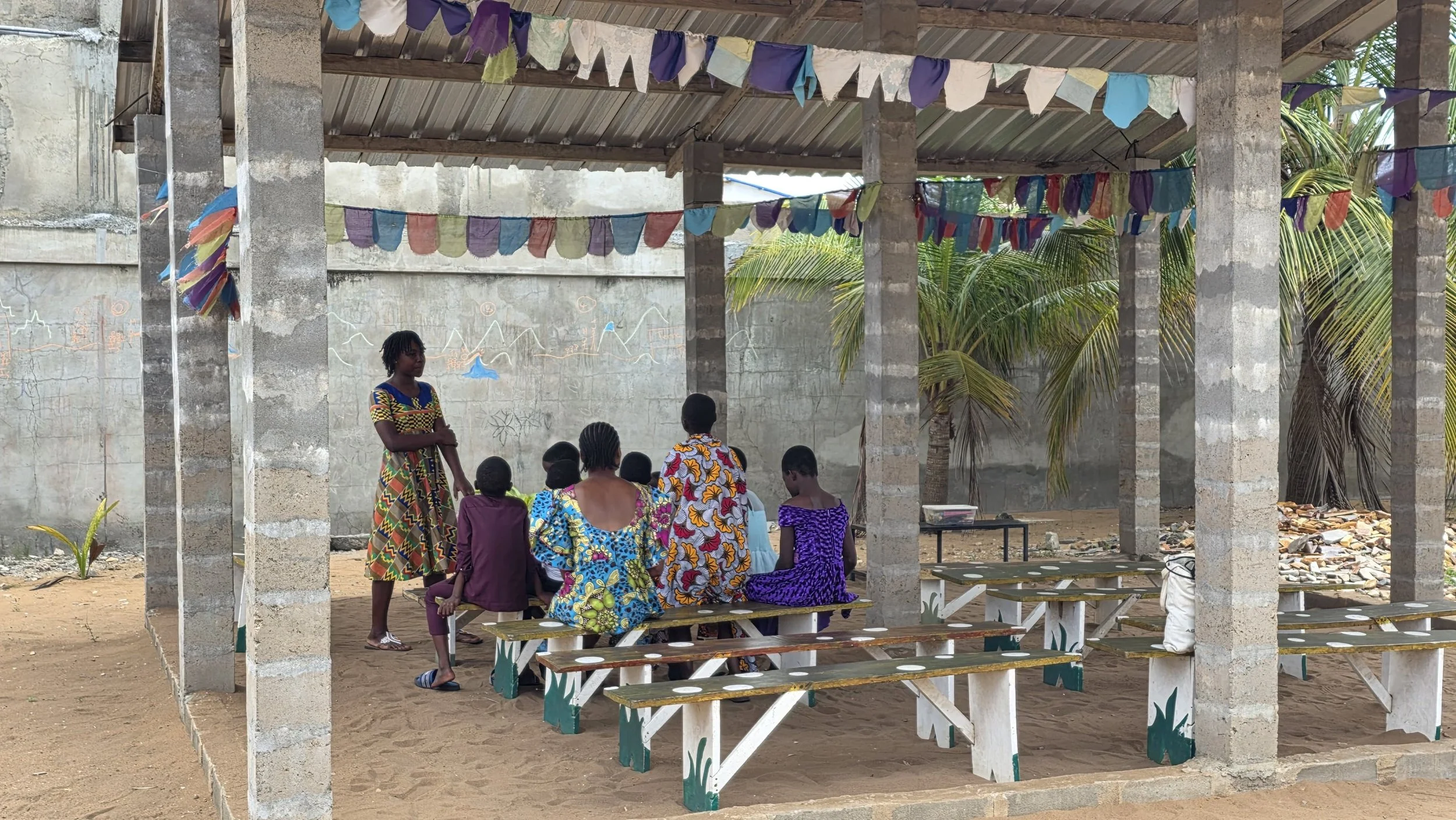 A group of women and children sitting on benches under a partially constructed shelter, with a woman standing and speaking to them. The shelter has a metal roof, concrete pillars, and colorful fabric bunting hanging from the ceiling.