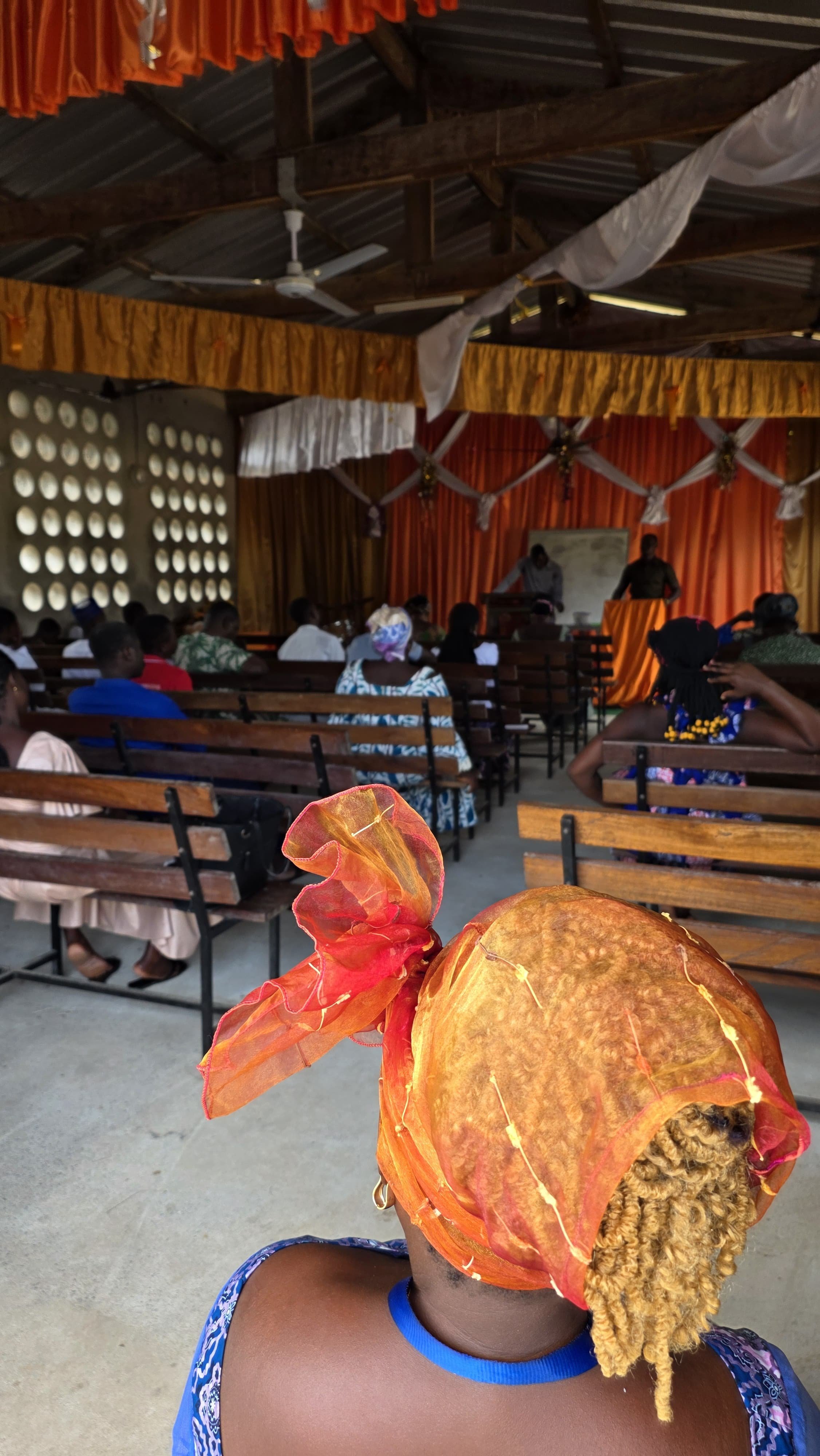 A woman wearing an orange head wrap with decorative yellow and orange accents, sitting in a hall with wooden benches, listening to two men at the front. The hall has orange and white draped curtains and a metal roof.