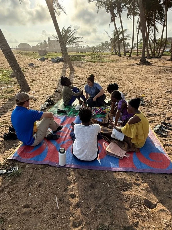 Group of children and two adults sitting on a beach towel under palm trees, reading and talking with each other, with the beach and ocean in the background.