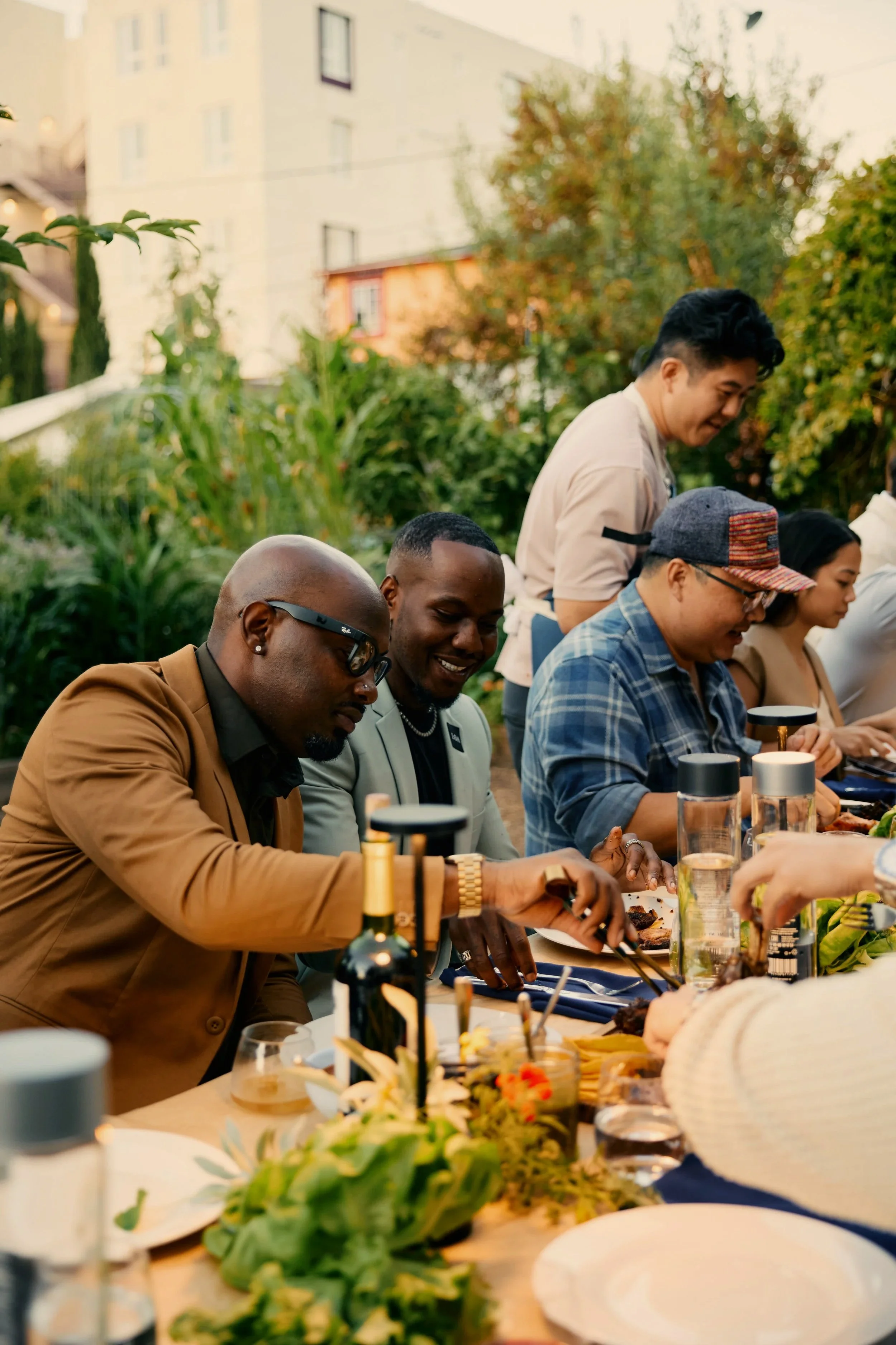 People enjoying a dinner outdoor gathering with food and drinks at a long table in a garden