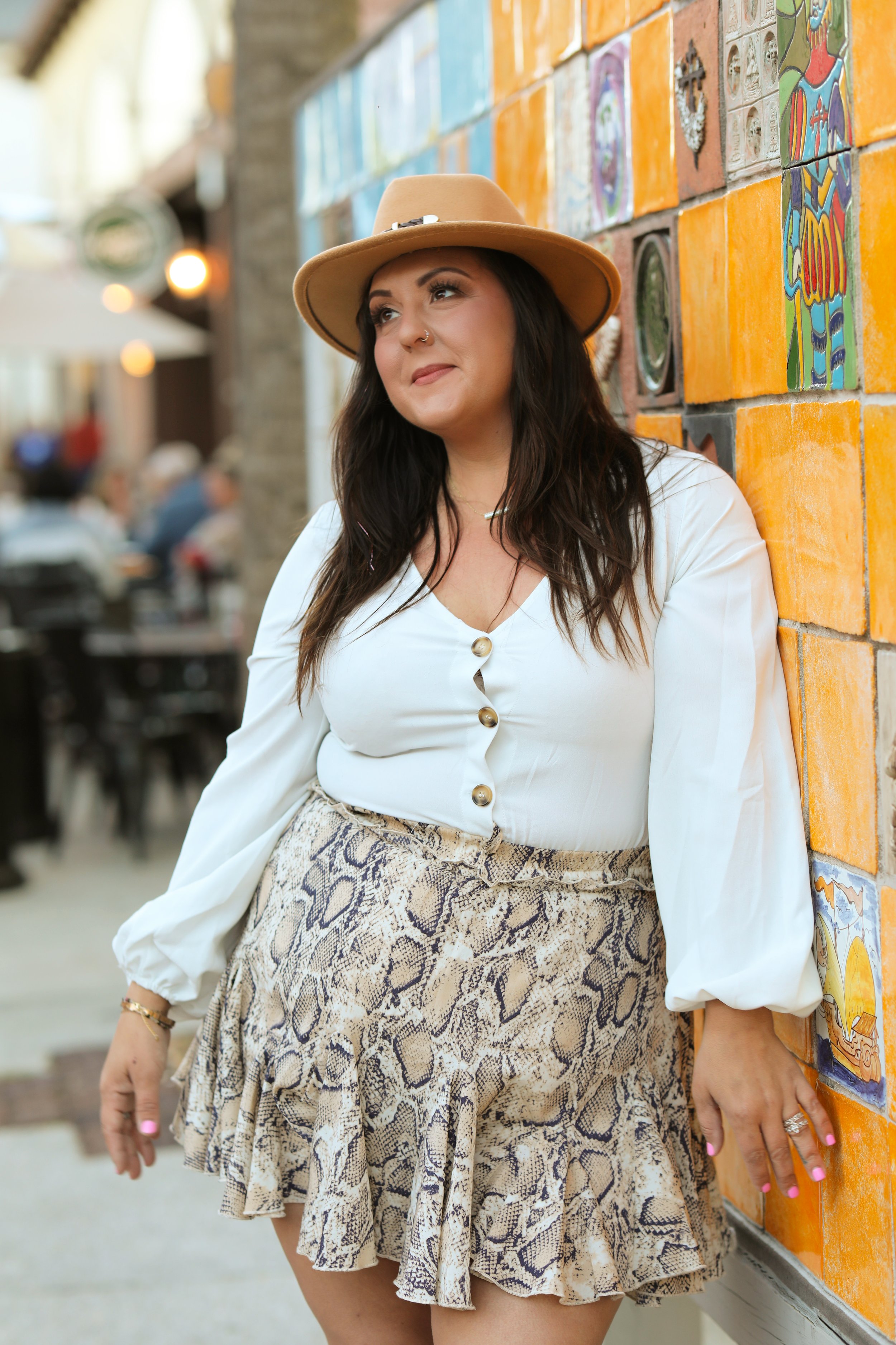 A woman with dark hair, wearing a beige hat, white blouse with buttons, and a snake print skirt, leaning against a colorful brick wall.