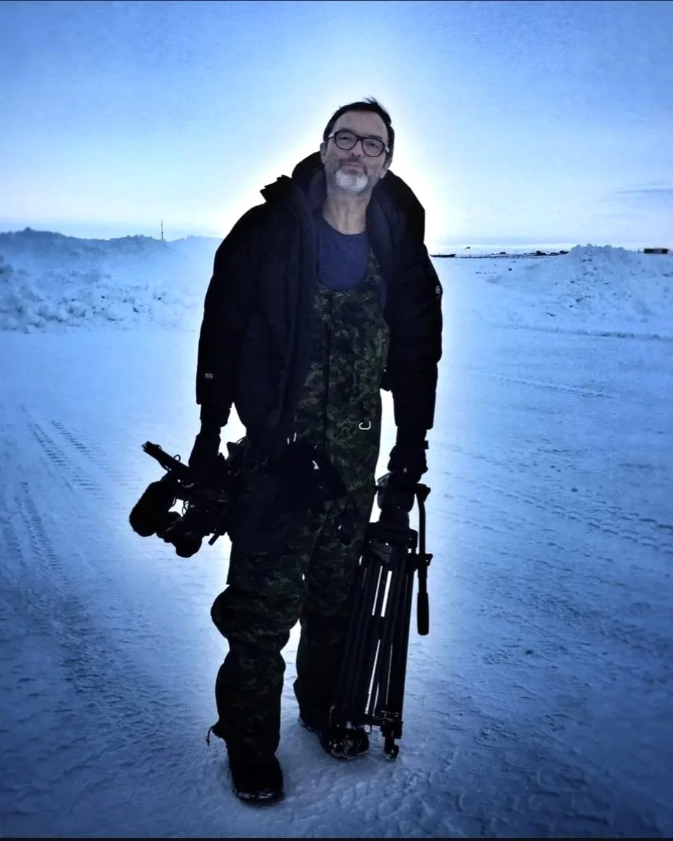 A man dressed in winter gear, with a camera and tripod, standing on a snowy landscape with ice and snow-covered ground at dusk or dawn.
