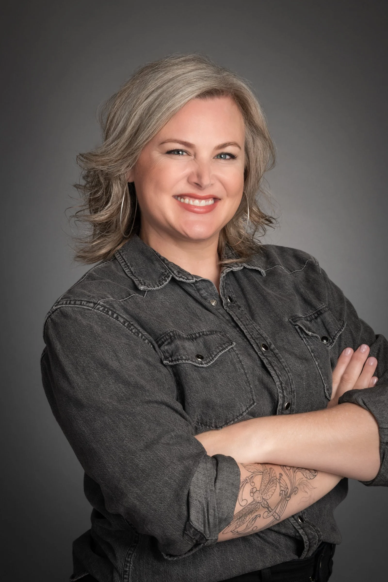A woman with shoulder-length blonde hair wearing a black denim shirt, smiling and crossing her arms, showing a tattoo on her left forearm, against a gray background.