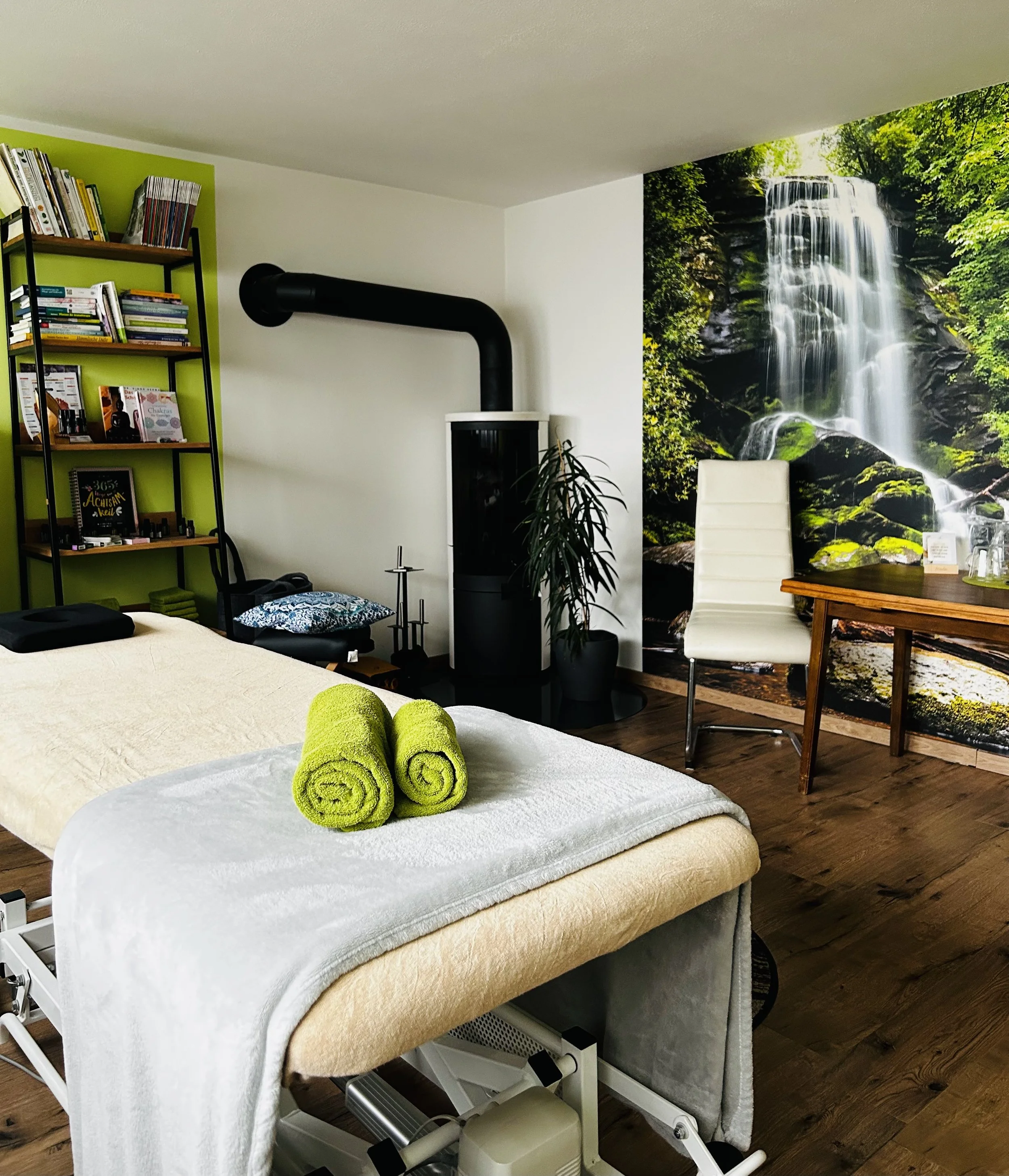 Ein Massagebett im Raum mit zwei grünen Handtüchern darauf, eine große Wasserfall-Wandtapete im Hintergrund und ein Bücherregal an der linken Wand.