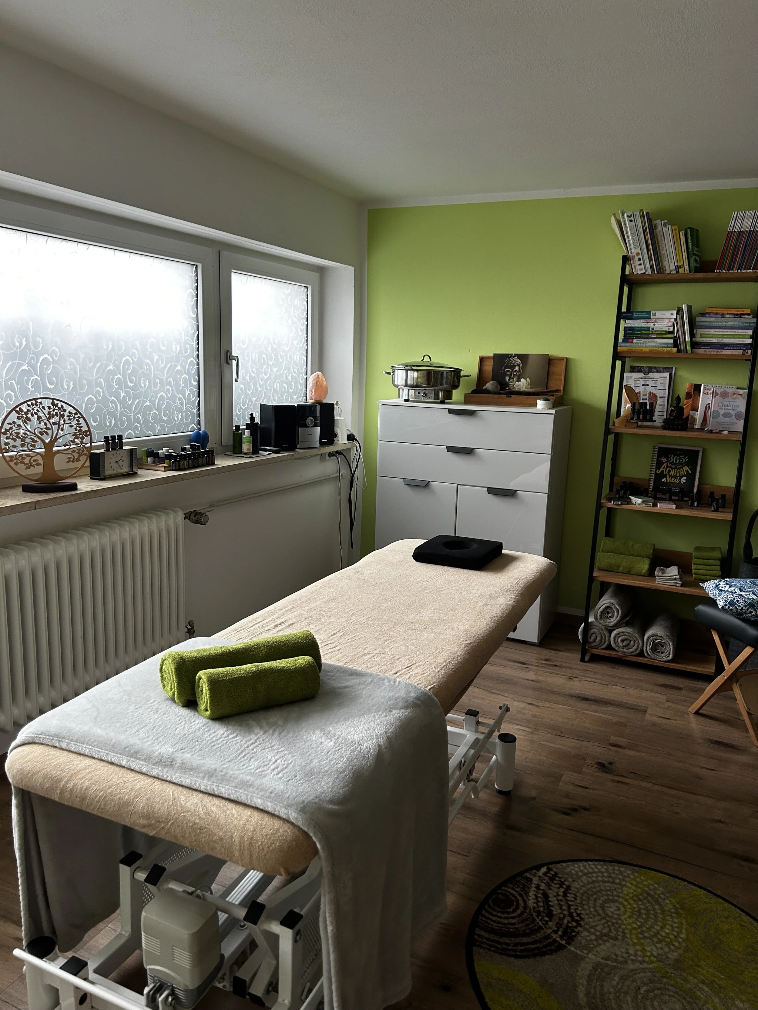 Behandlungsraum mit Massagebett, grüner Wand, Regal mit Büchern und Wellness-Tools, Fenster mit dekorativen Vorhängen, Holzfußboden.