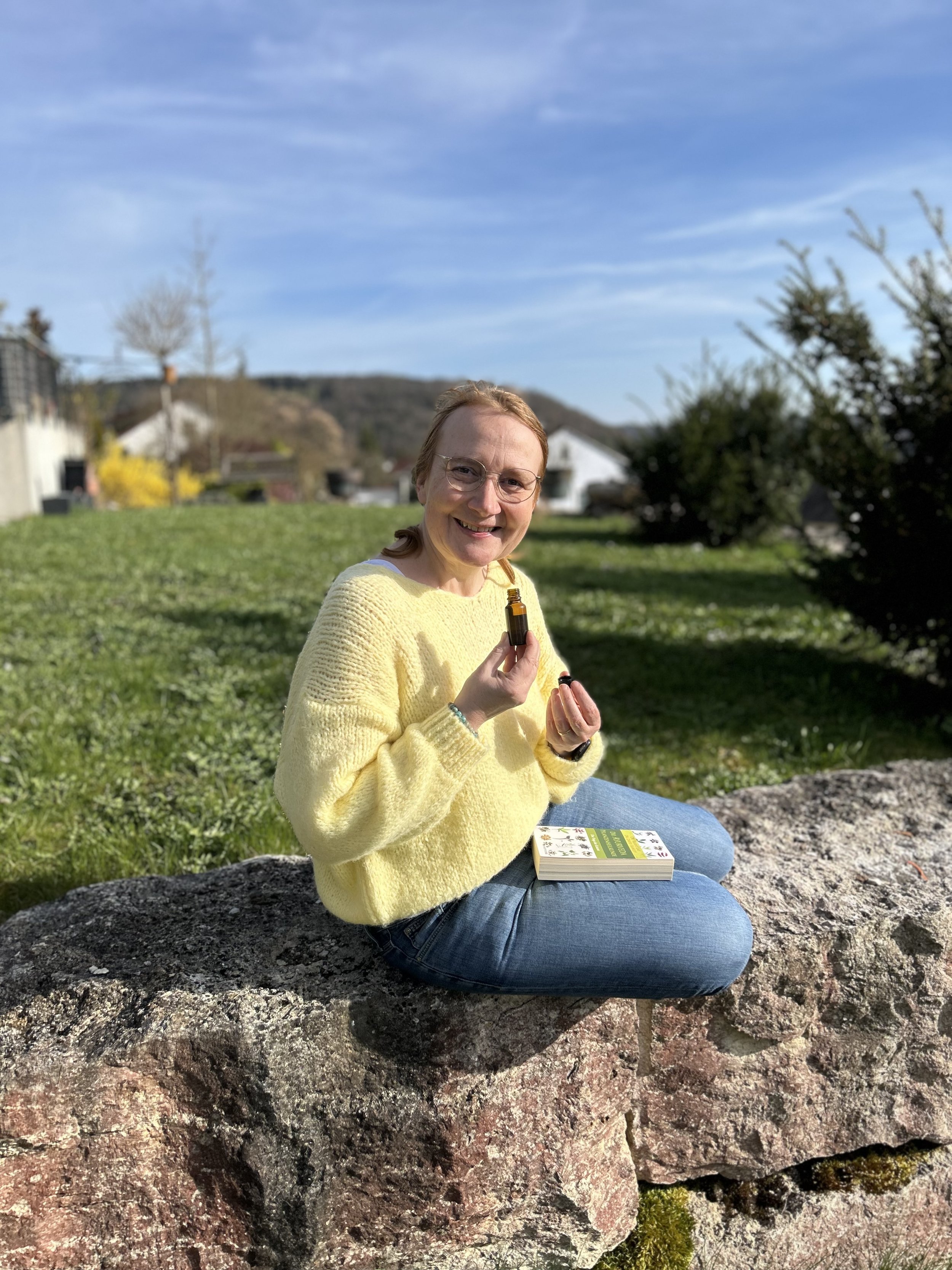 Eine Frau in einem gelben Pullover sitzt auf einem Felsen im Freien, hält eine kleine Flasche in der Hand und hat ein Buch auf den Knien, während sie in die Kamera lächelt.
