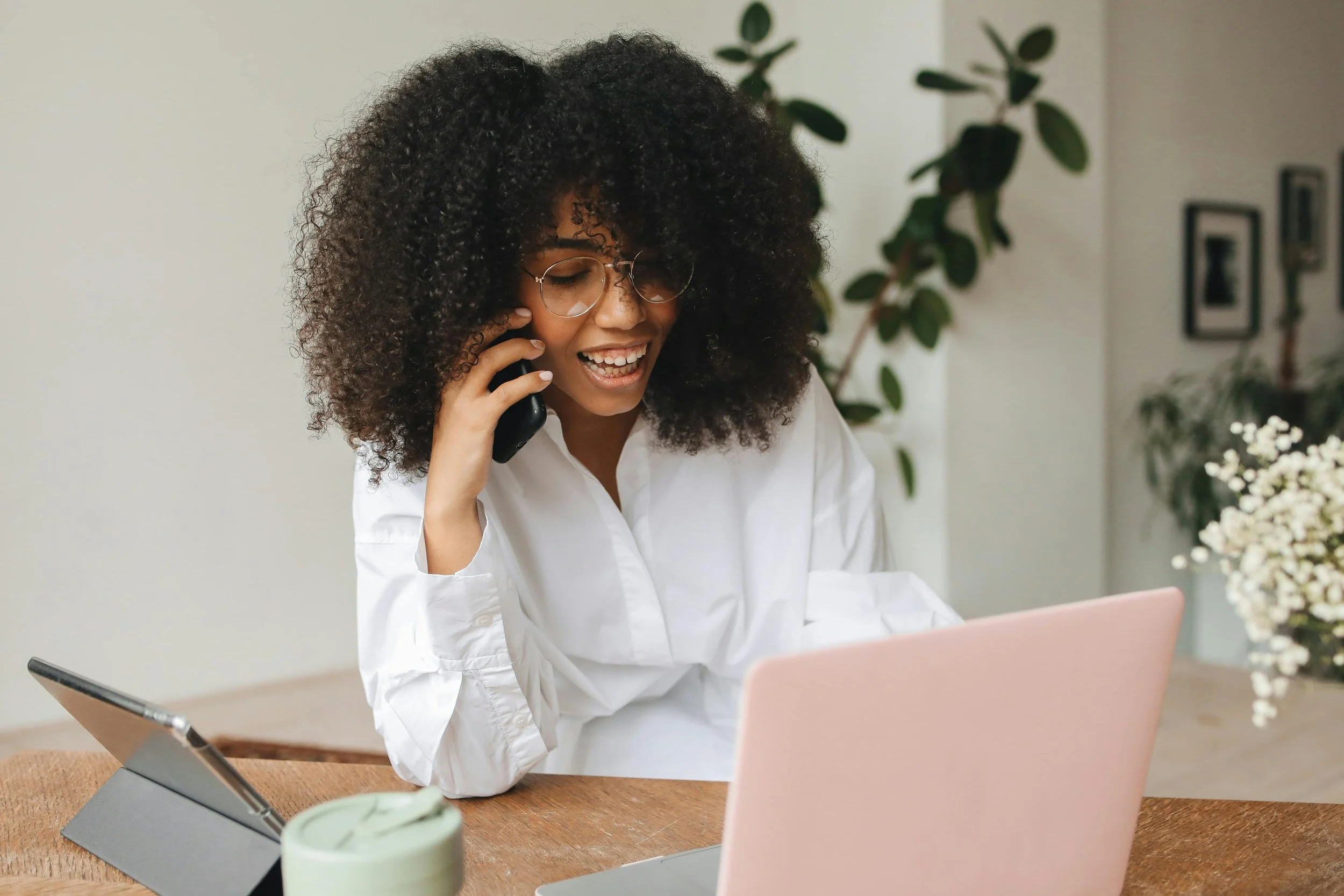 A woman with curly hair, glasses, and a white shirt sits at a wooden table with a pink laptop, talking on a phone, smiling, in a room with indoor plants and framed pictures.