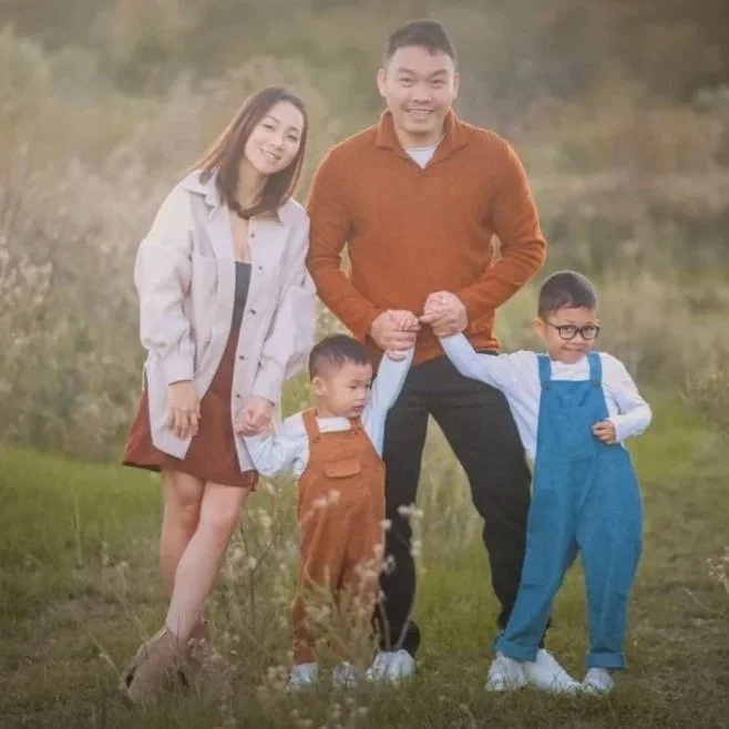 Family of four standing outdoors on grass, holding hands and smiling; the mother wears a white jacket and brown dress, father wears an orange sweater, with two children dressed in overalls, one in brown and the other in blue.
