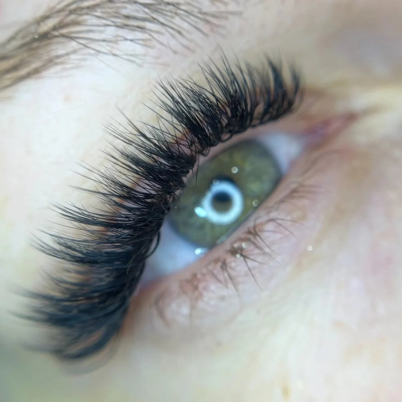 Close-up of a human eye with long, thick eyelashes and a green iris, reflecting a ring light.