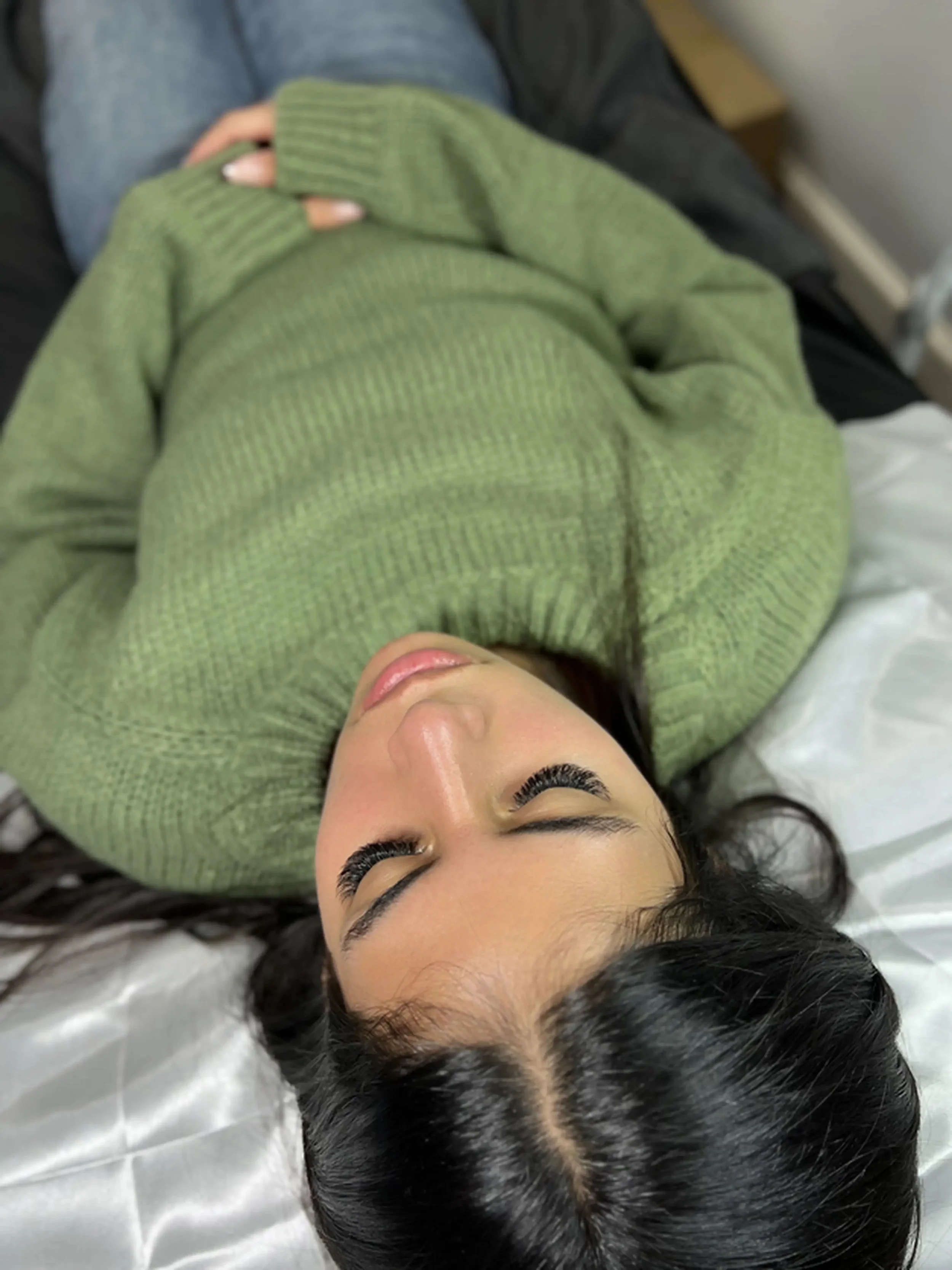 a woman lying on their back wearing a green sweater getting a new set of lash extension done.