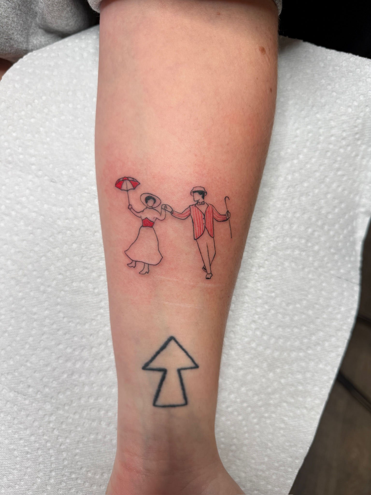A tattoo of a line art illustration of a man and woman holding hands and dancing, with the woman holding a red umbrella. The man is wearing a hat, and the woman has a wide-brimmed hat. There is a black arrow pointing upwards below the dancing couple.