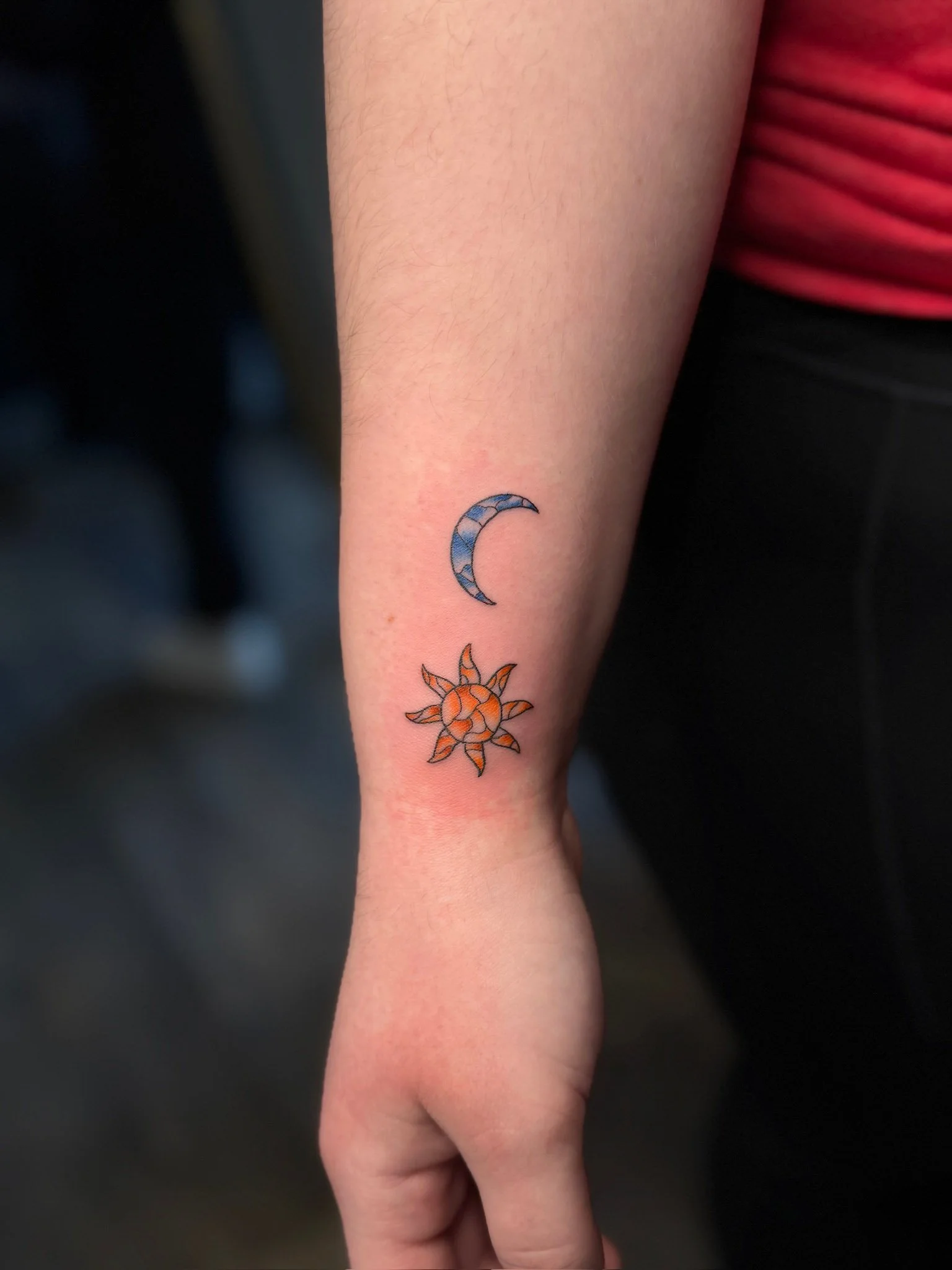 Tattoo of a blue crescent moon above an orange sun on person's inner forearm.
