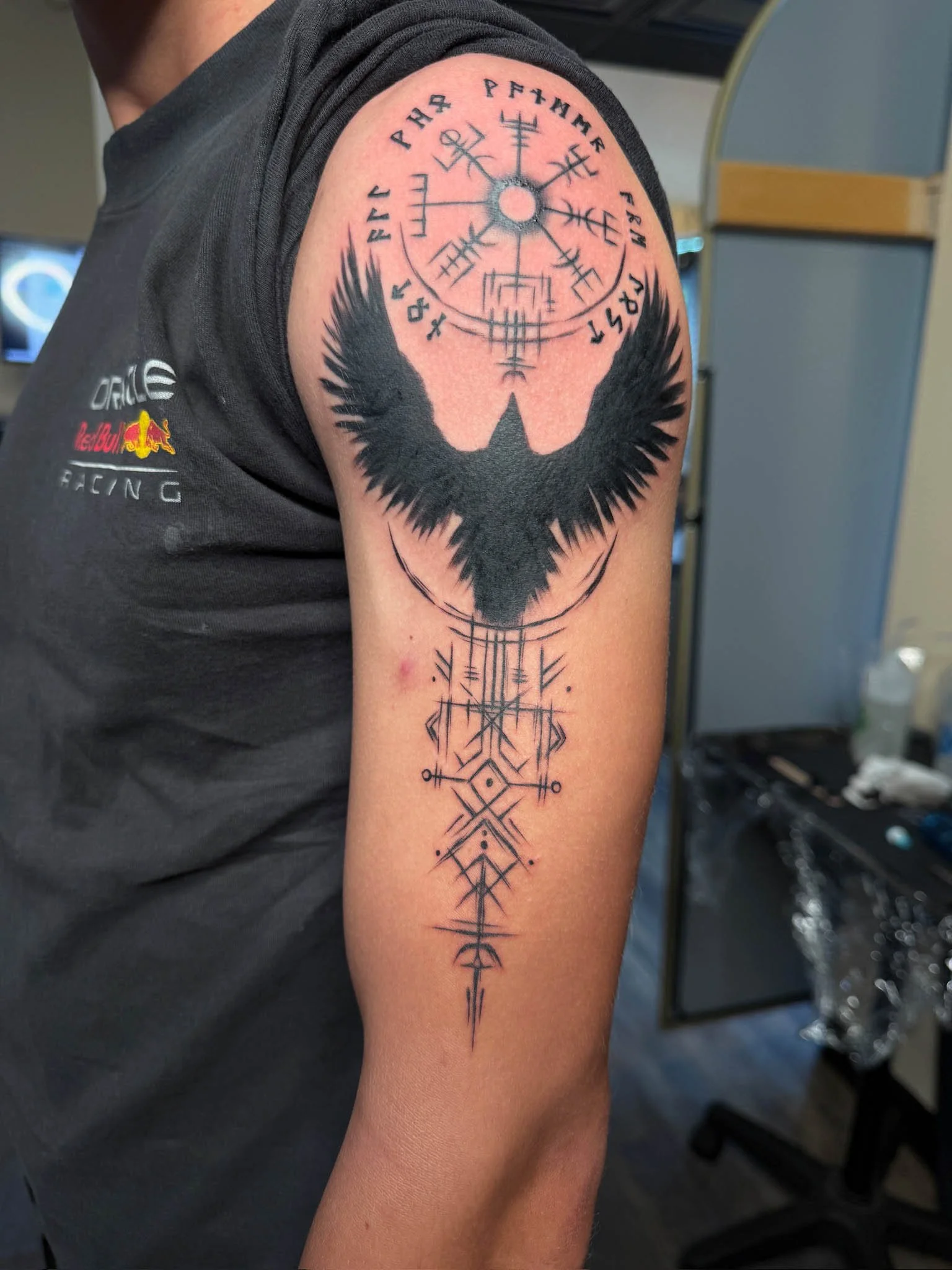 Tattoo on person's upper arm featuring a black bird with outstretched wings and a circular Nordic-style symbol above it, with geometric lines and shapes extending downward.
