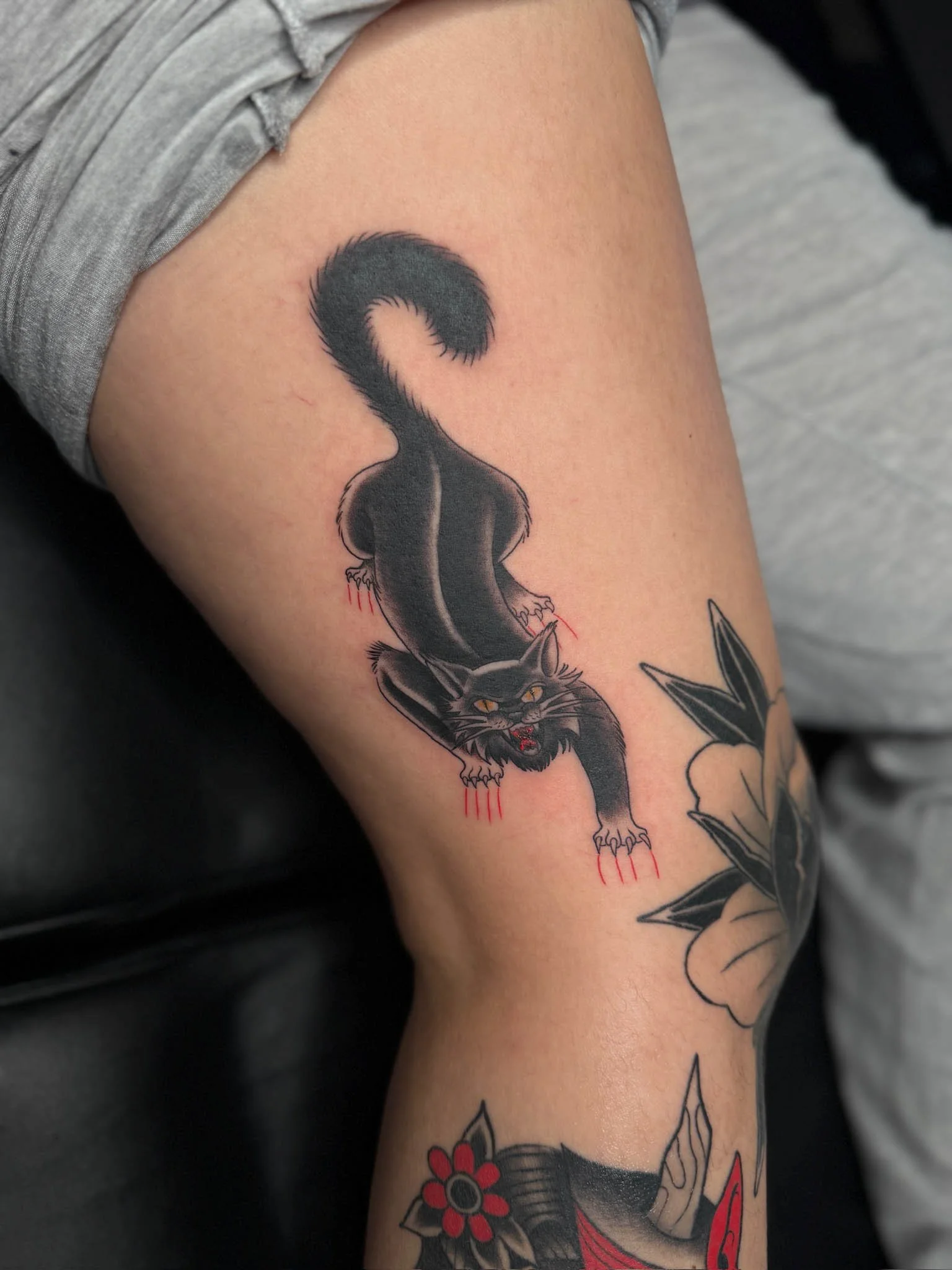 Tattoo of a black cat with yellow eyes, showing claws with red accents, on a person's upper arm.