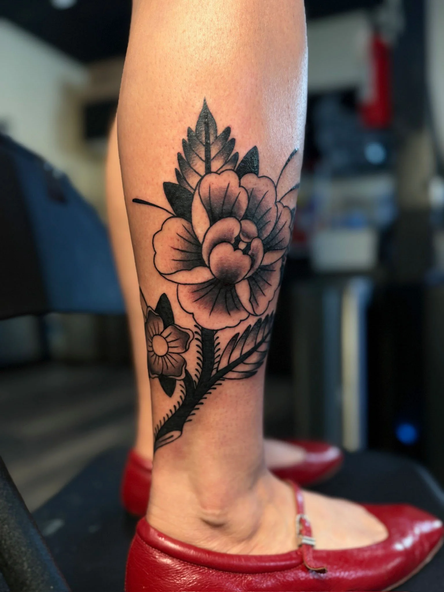 A tattoo of a large flower with surrounding leaves on a person's lower leg, with another smaller flower below it. The person is wearing red shoes.