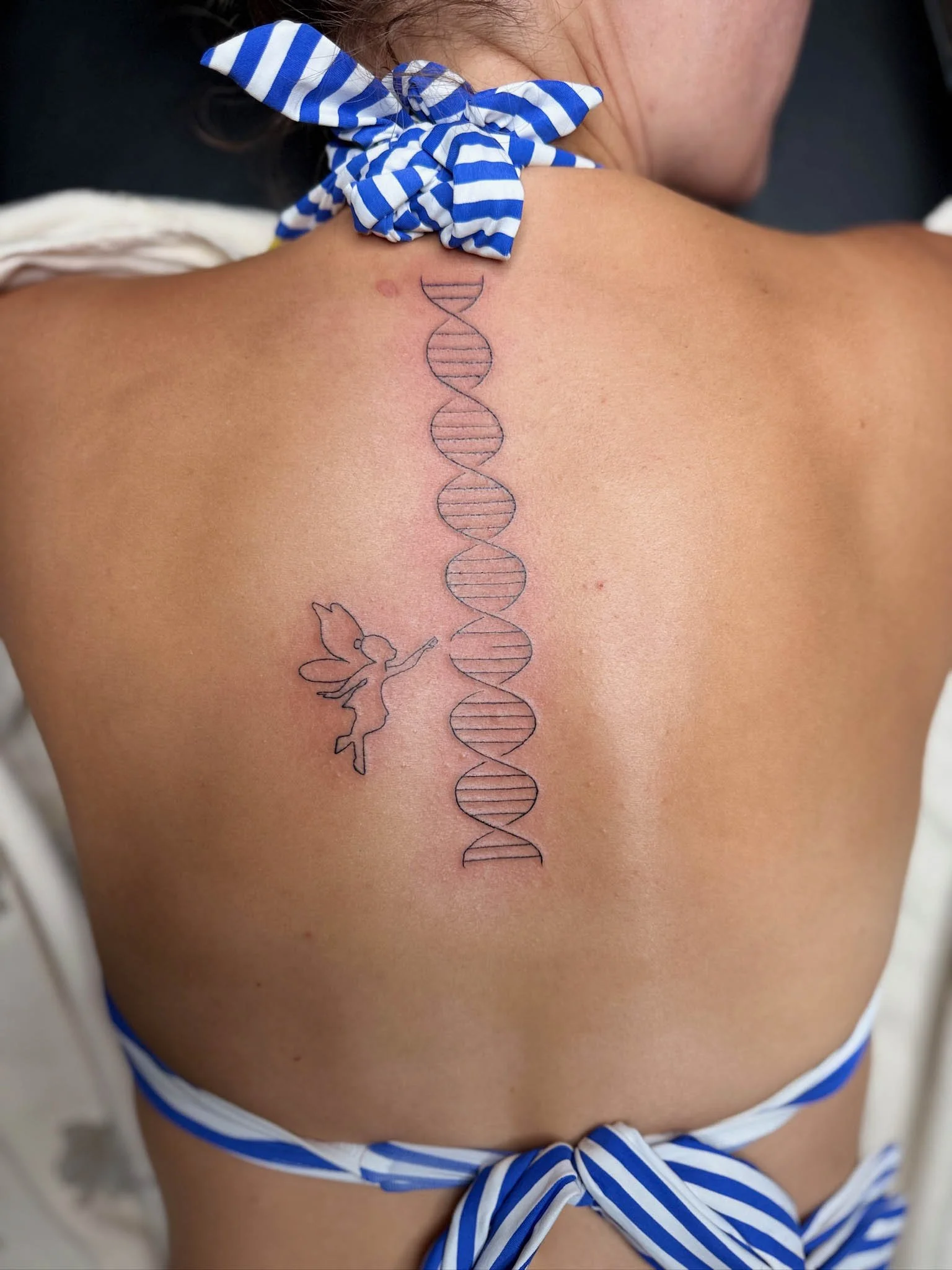 Tattoo of a DNA double helix with a fairy reaching out to it on a person's back.
