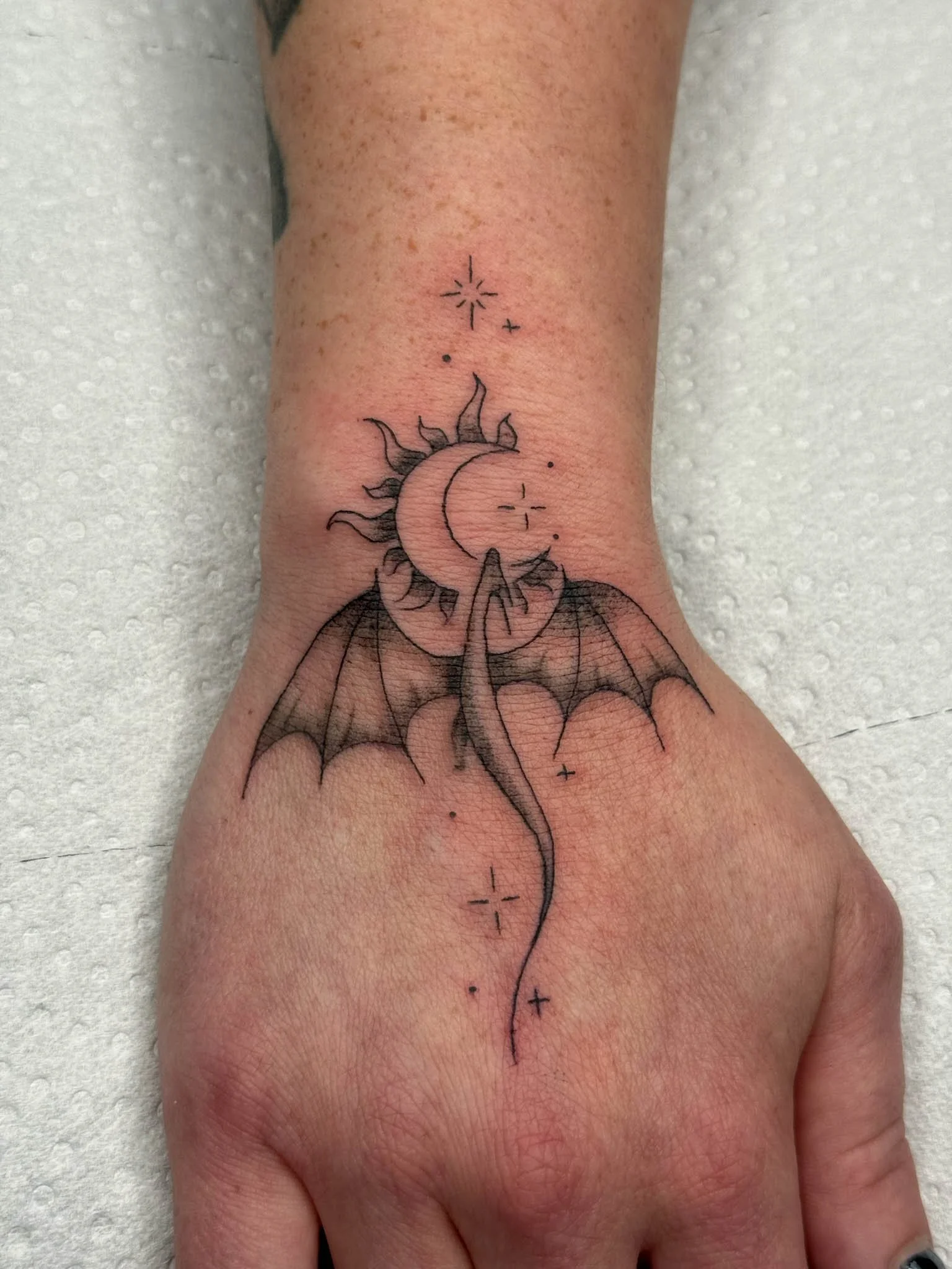 Tattoo of a moon with bat wings, a sun surrounded by stars, and a long tail on a wrist.