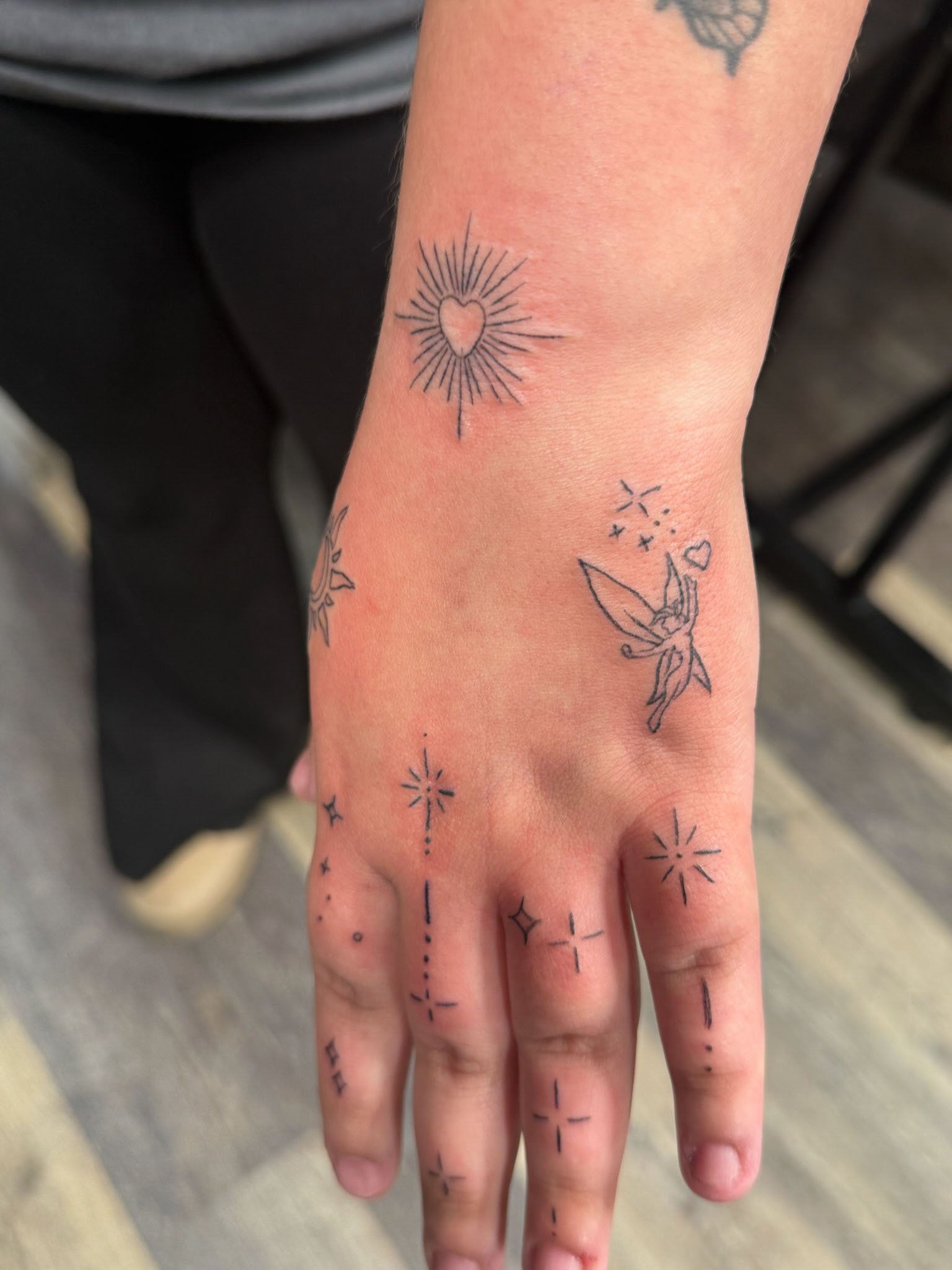 A person's hand and wrist with multiple small tattoo designs, including a heart surrounded by rays, a fairy with wings, and various star and line patterns.