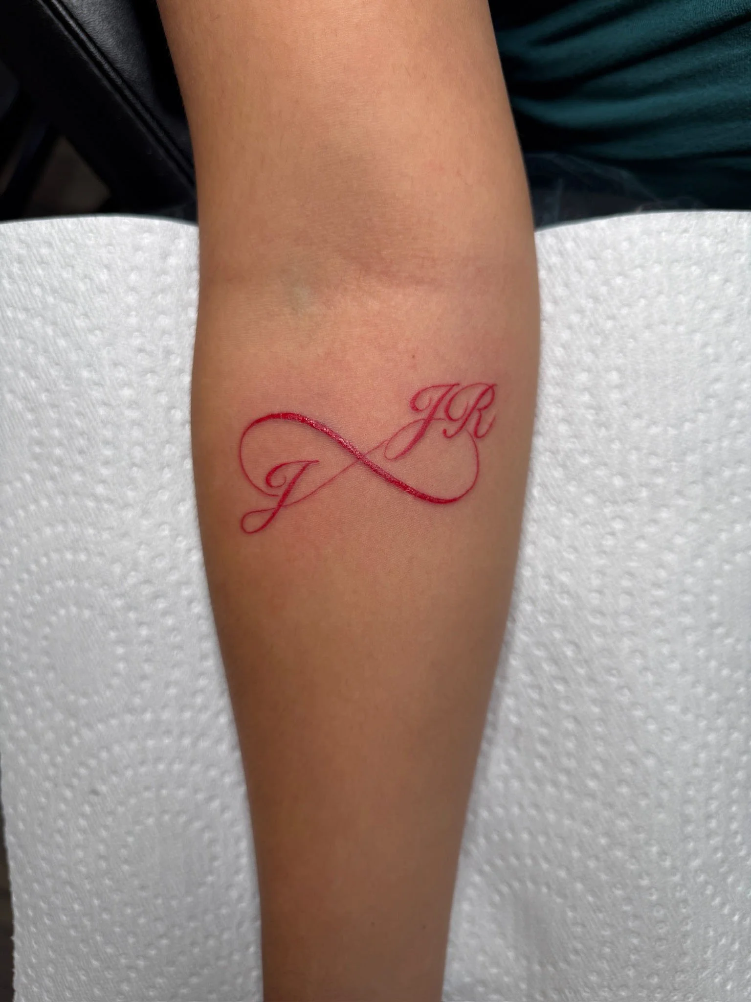 Tattoo of the initials J R in red ink on a person's forearm, with the initials connected by an infinity symbol.