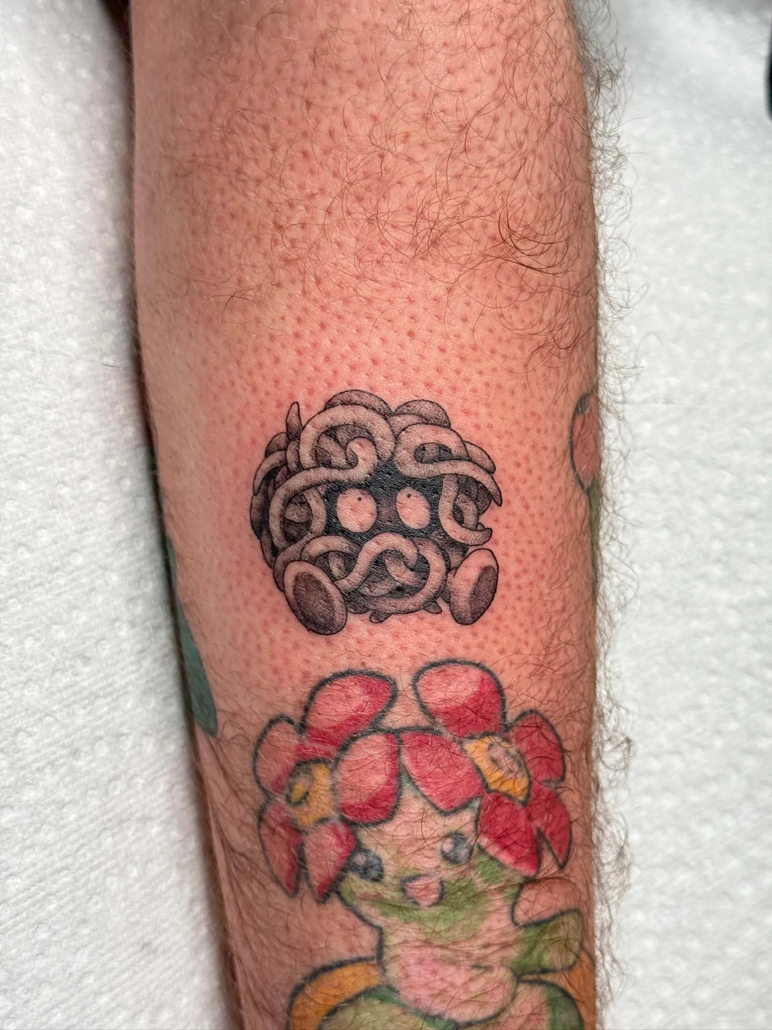 Tattoo of a creature made of noodles with two eyes, above a drawing of a girl with a red flower hat and long hair.