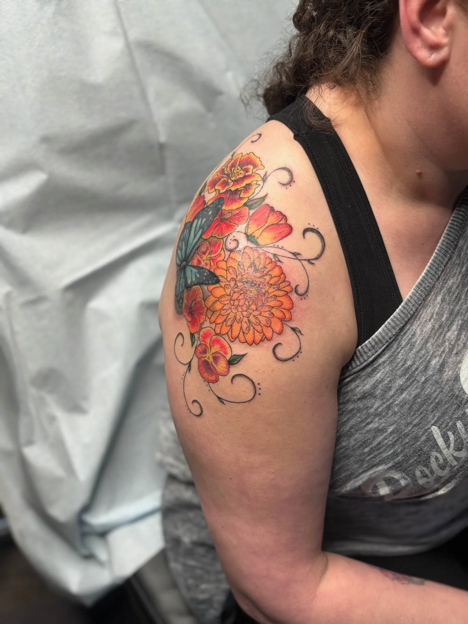 Colorful floral tattoo with orange flowers and a blue butterfly on a person's shoulder.