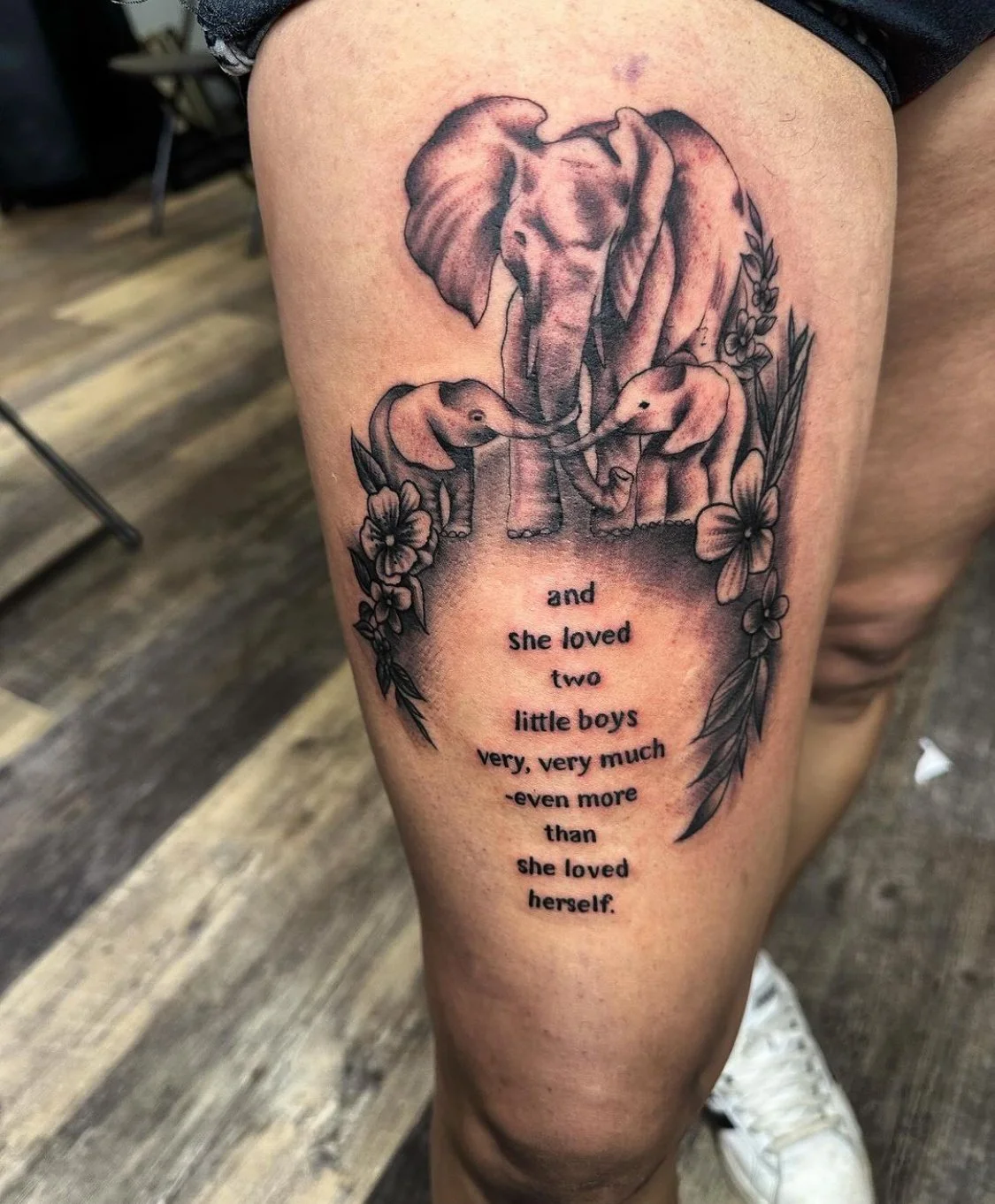 Close-up photograph of a black and grey realism tattoo showing an elephant family and a quote on thigh