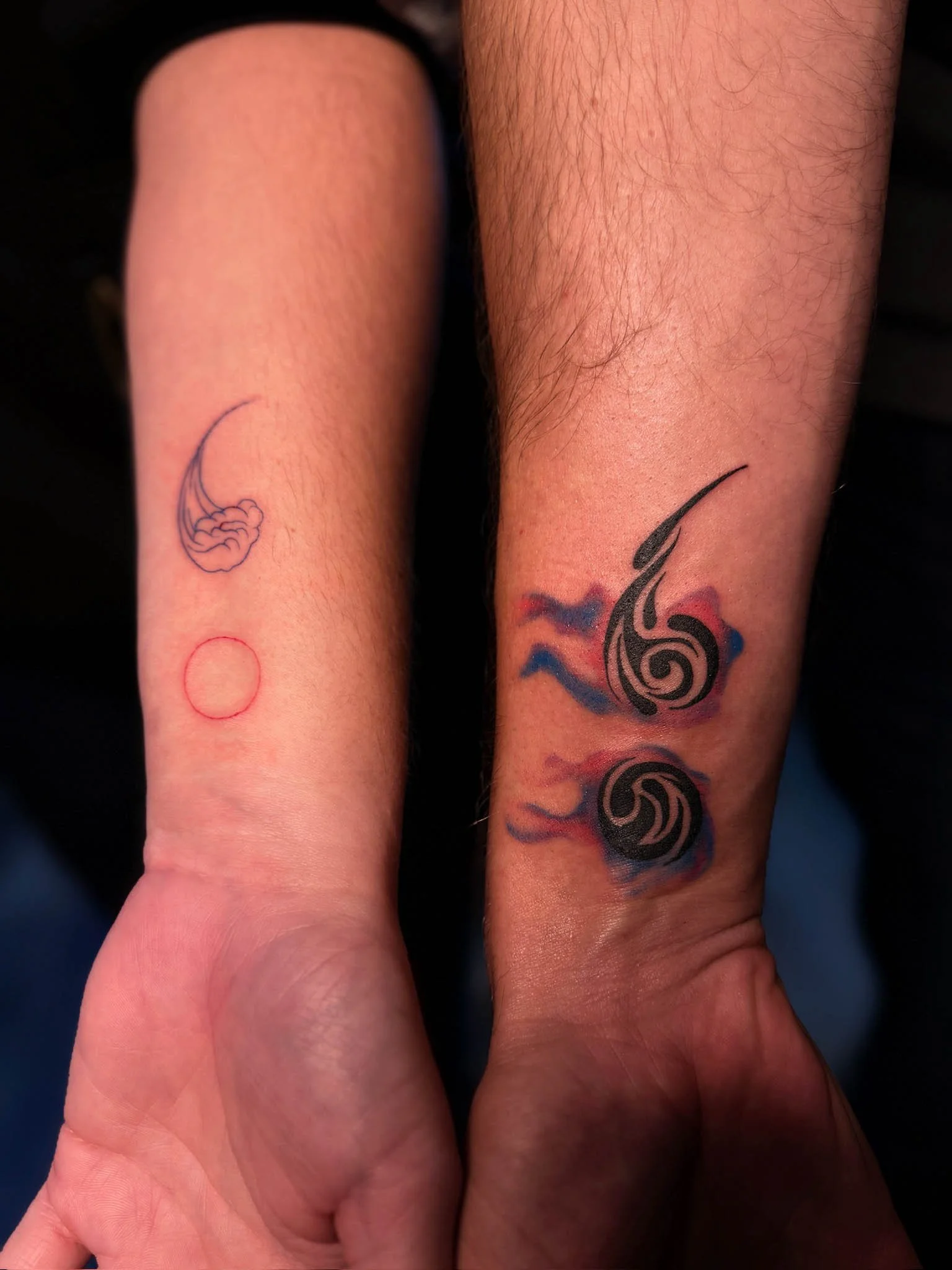 Two people's forearms with tattoos. The arm on the left has a small, simple tattoo of a claw and an outline of a circle. The arm on the right has a larger, colorful tattoo of two black gnarled spirals with red and blue smoke-like accents.