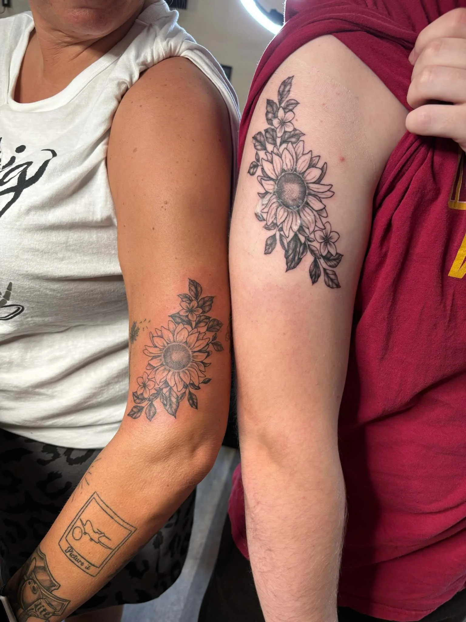 Two people show off their upper arms with matching sunflower tattoos on their biceps.