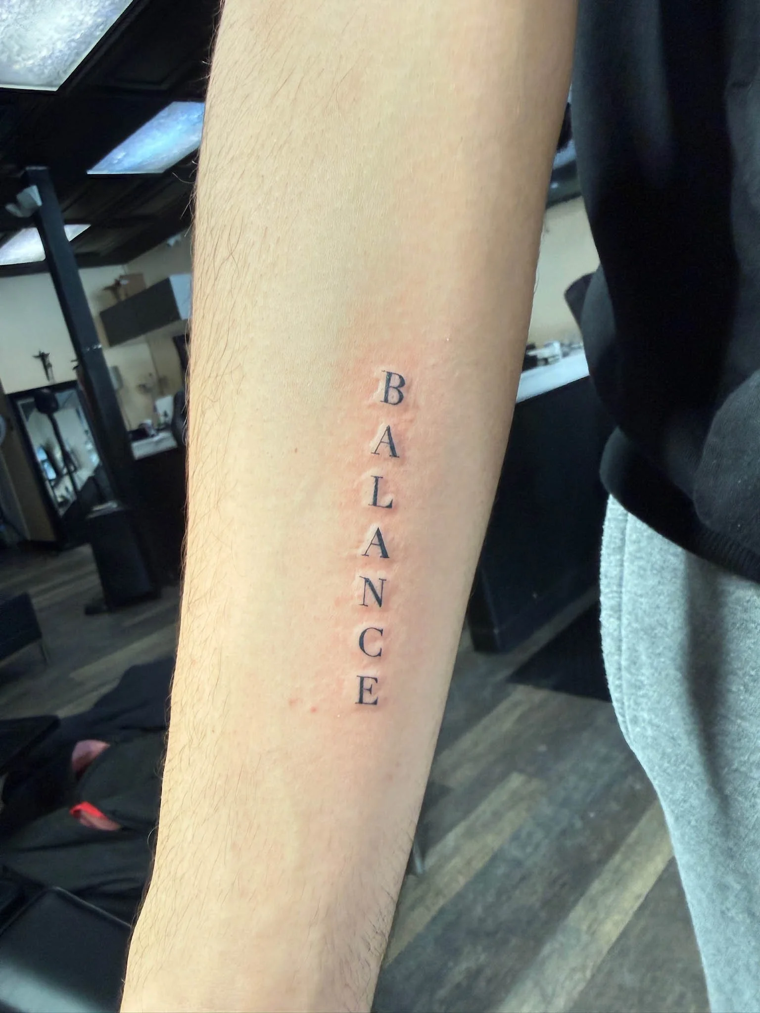 A fresh tattoo on a person's forearm with the word 'BALANCE' written vertically in black ink.