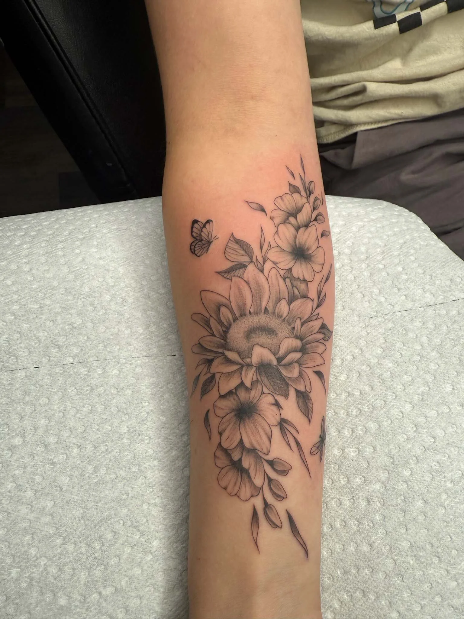 A tattoo of a sunflower with a butterfly near a bunch of other flowers and leaves on a person's forearm.