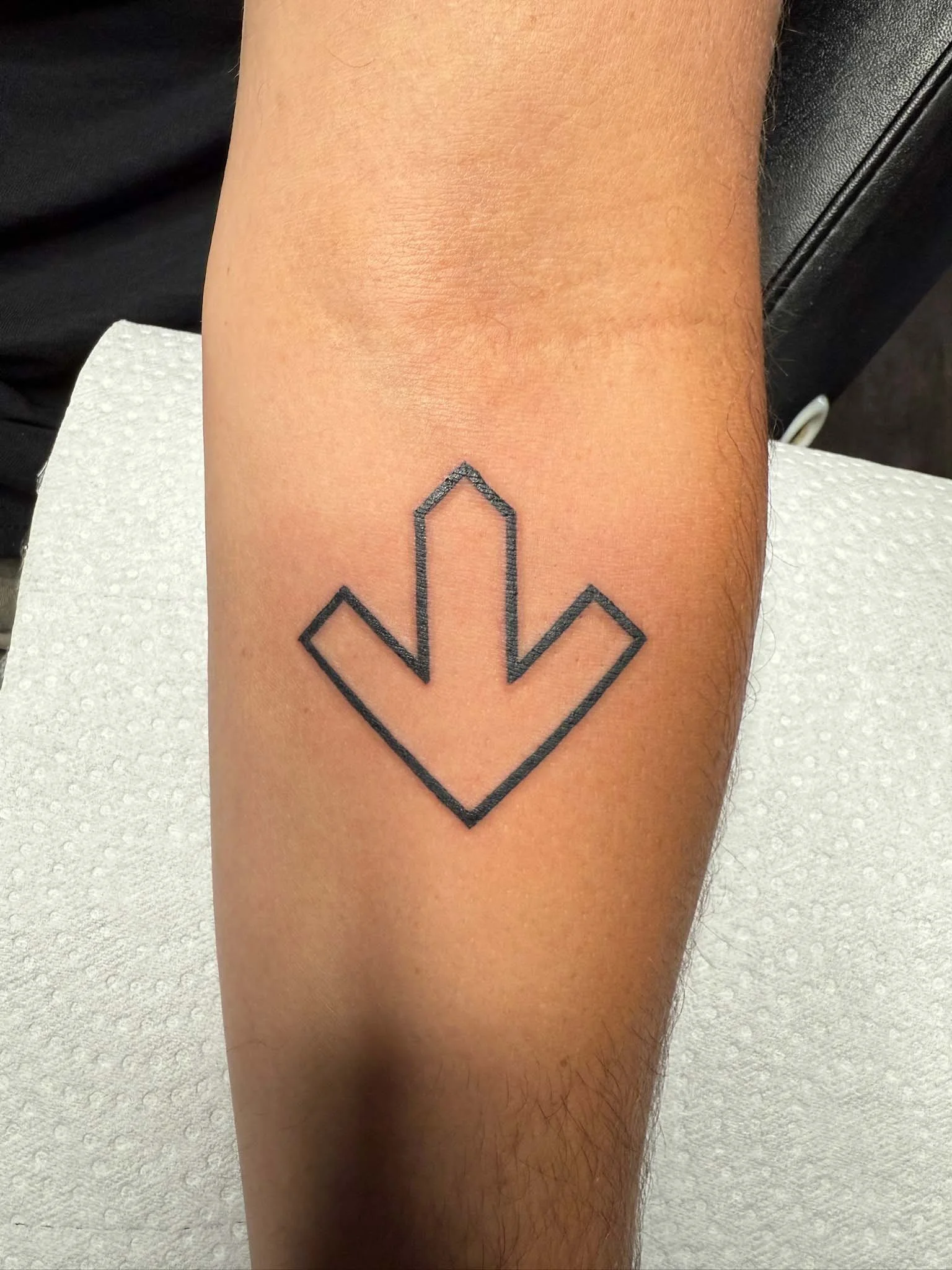Tattoo of a black outline downward arrow on a person's forearm.