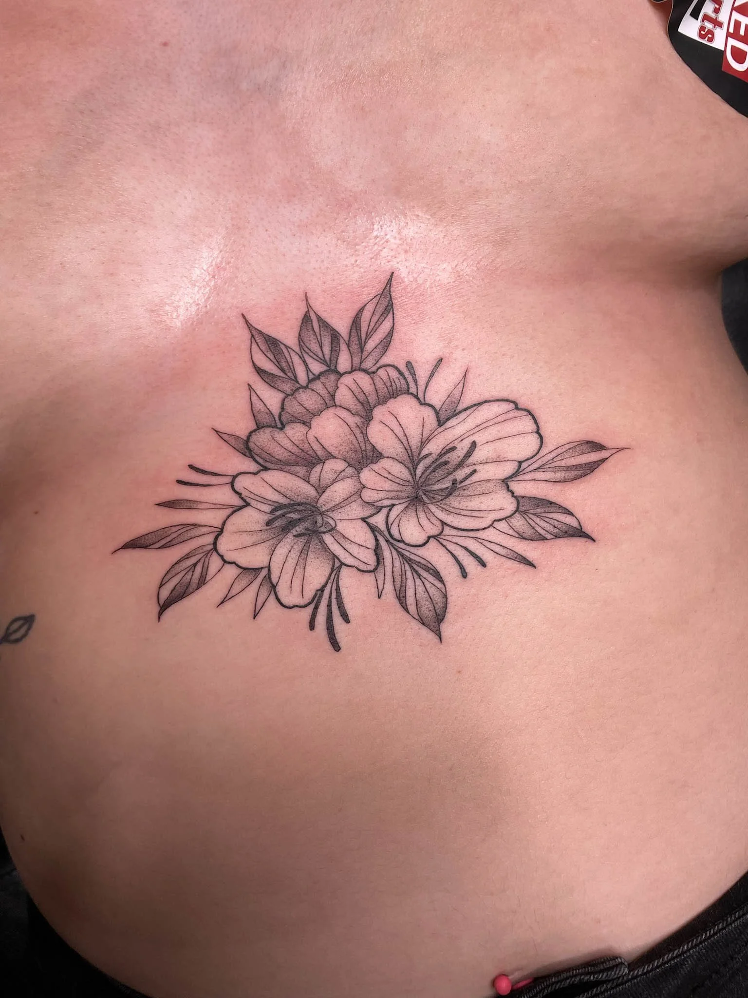 A black and gray floral tattoo featuring two large flowers with multiple petals and several leaves on a person's upper chest.