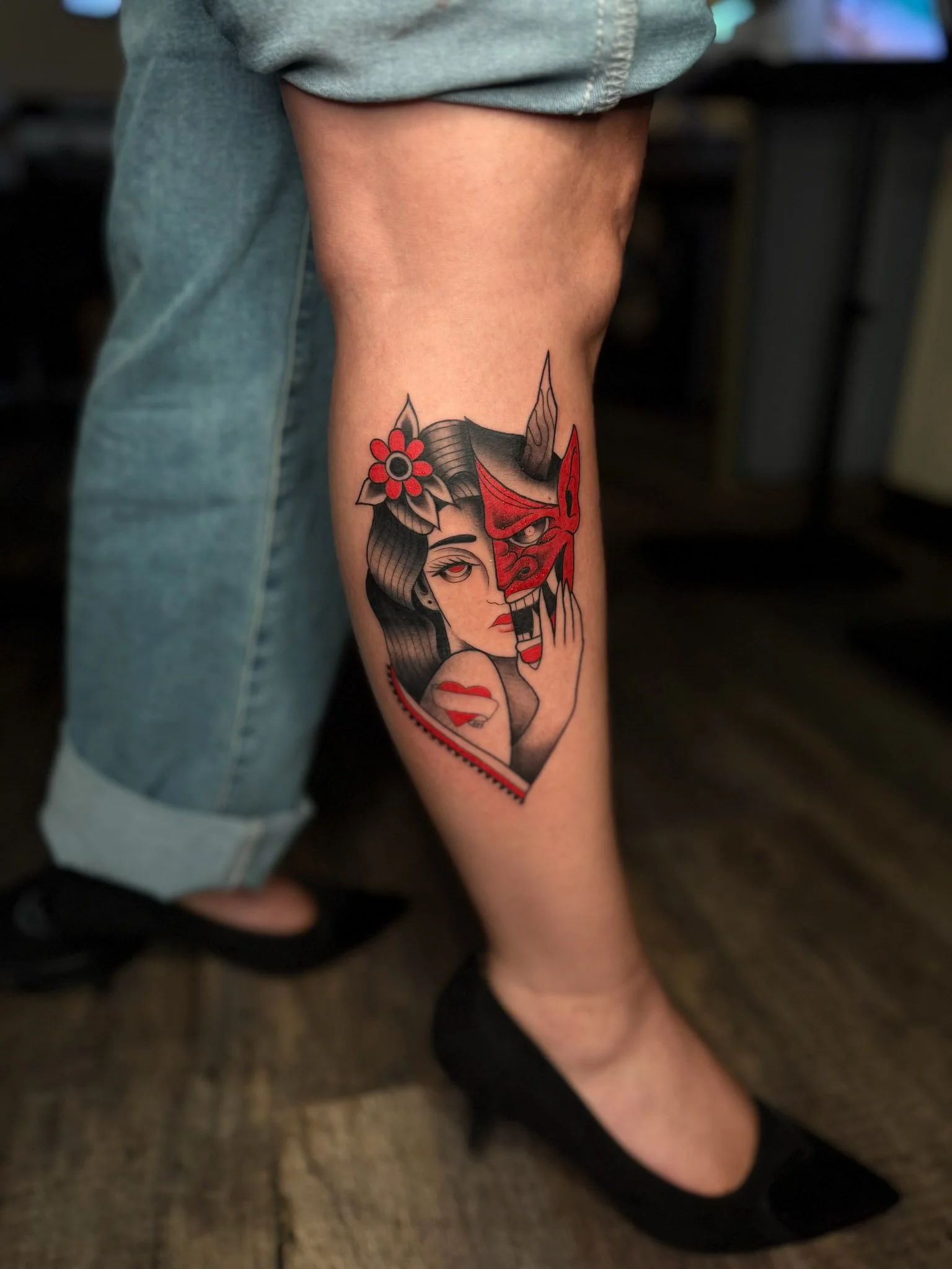 Tattoo of a woman with black hair, red lips, and a red flower in her hair, holding a red and black demon mask, on a person's calf.