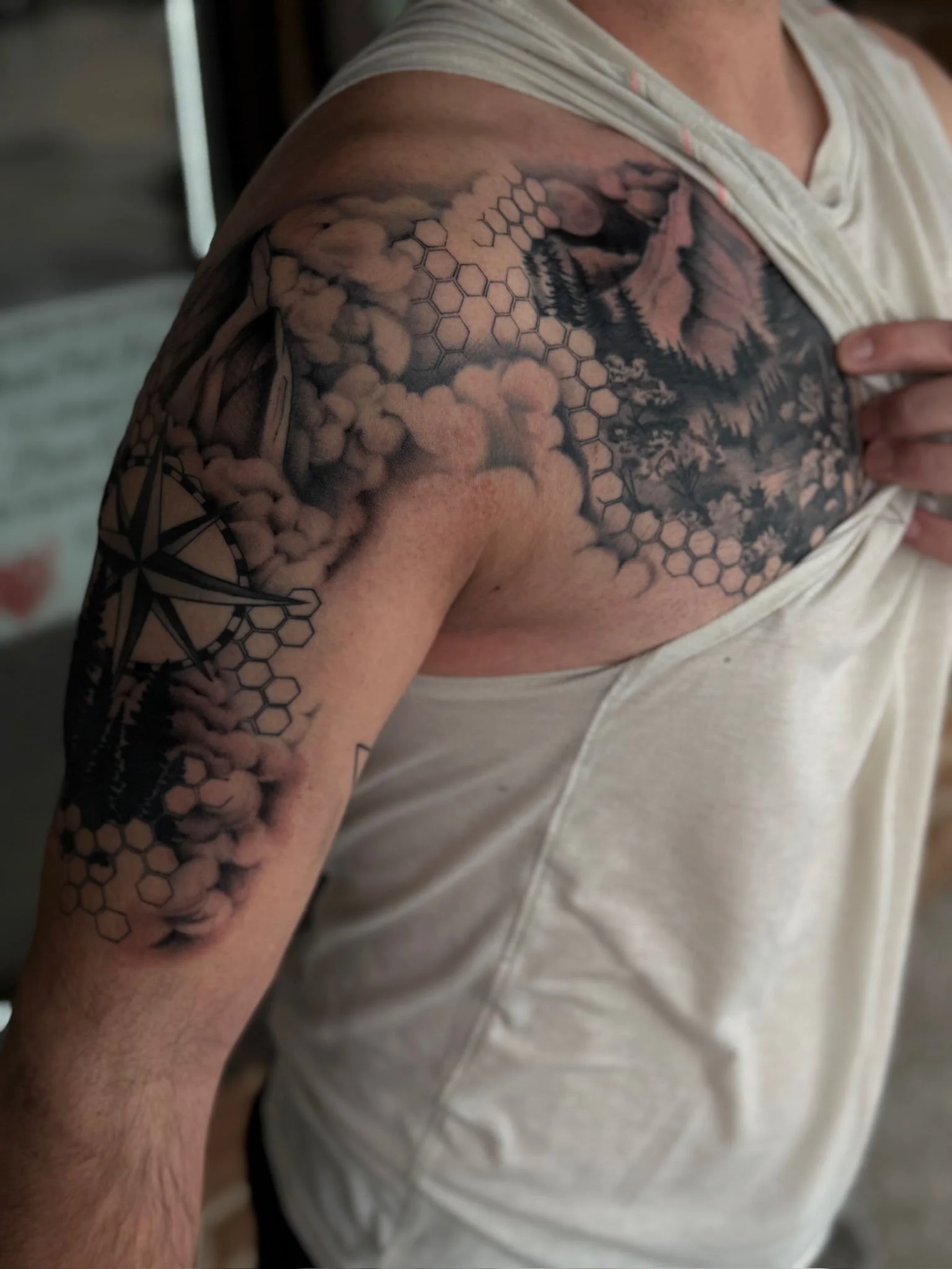 A man's shoulder and chest with a tattoo featuring clouds, mountains, trees, honeycomb patterns, and a compass.