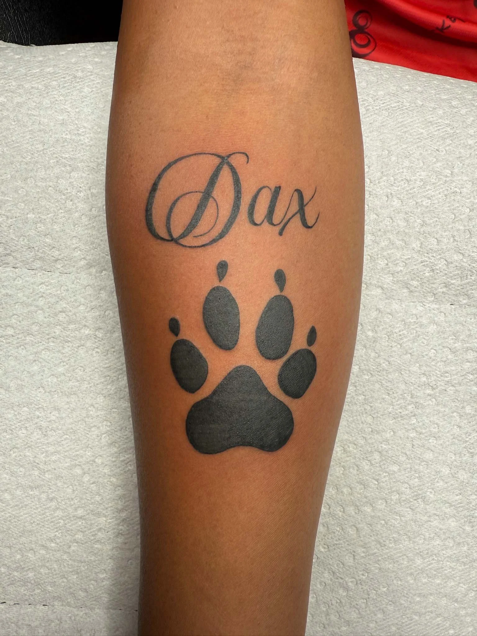 Tattoo of the word 'Dax' written in cursive above a black paw print on a person's forearm.