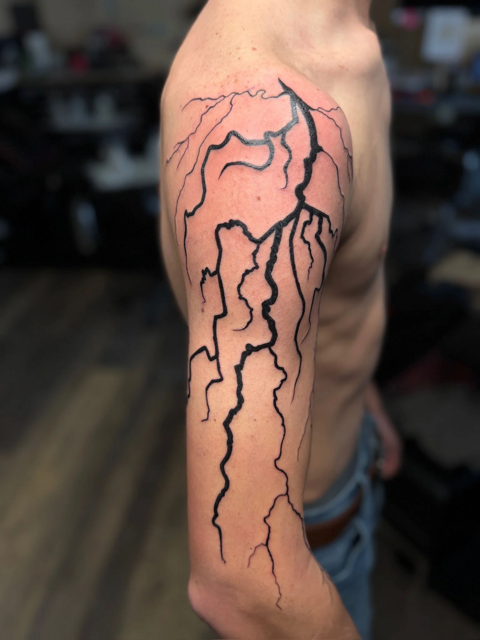 A person's upper arm with a tattoo of a network of black lightning bolts extending from the shoulder down the arm.