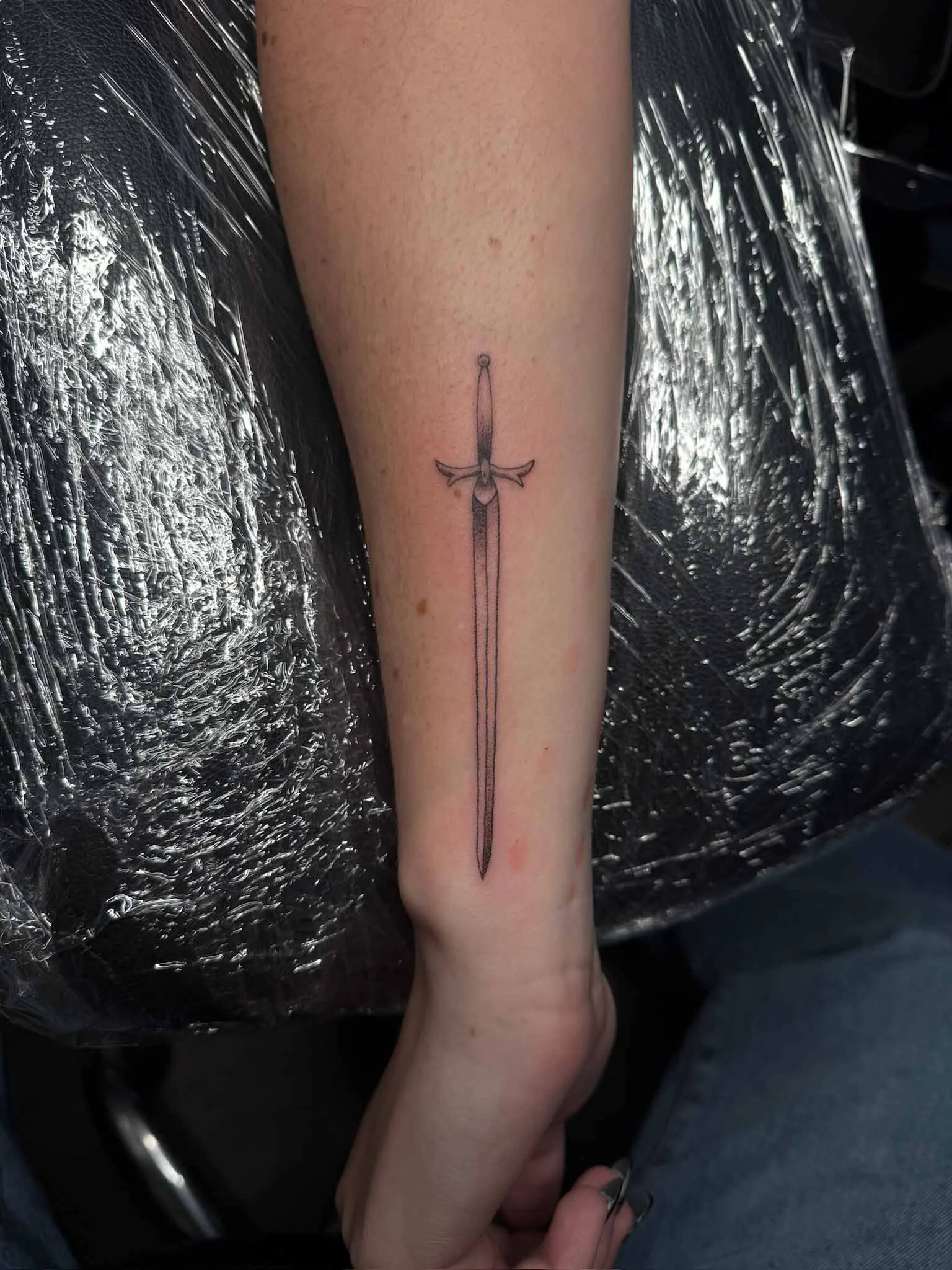 A tattoo of a sword on a person's forearm, with a simple line drawing style.