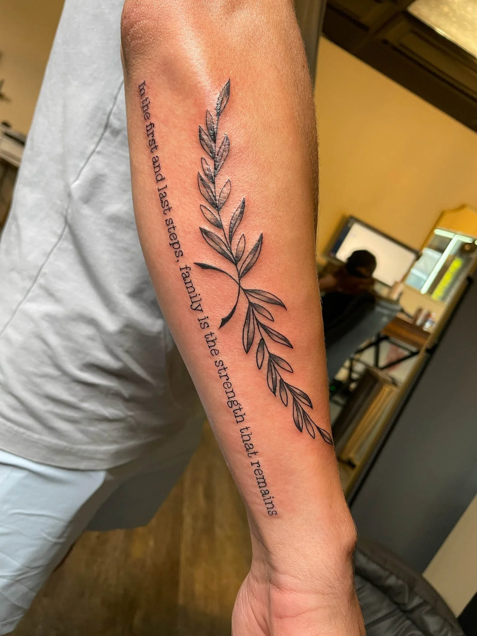 A tattoo of a leafy branch with a quote written vertically in black ink on a person's forearm.