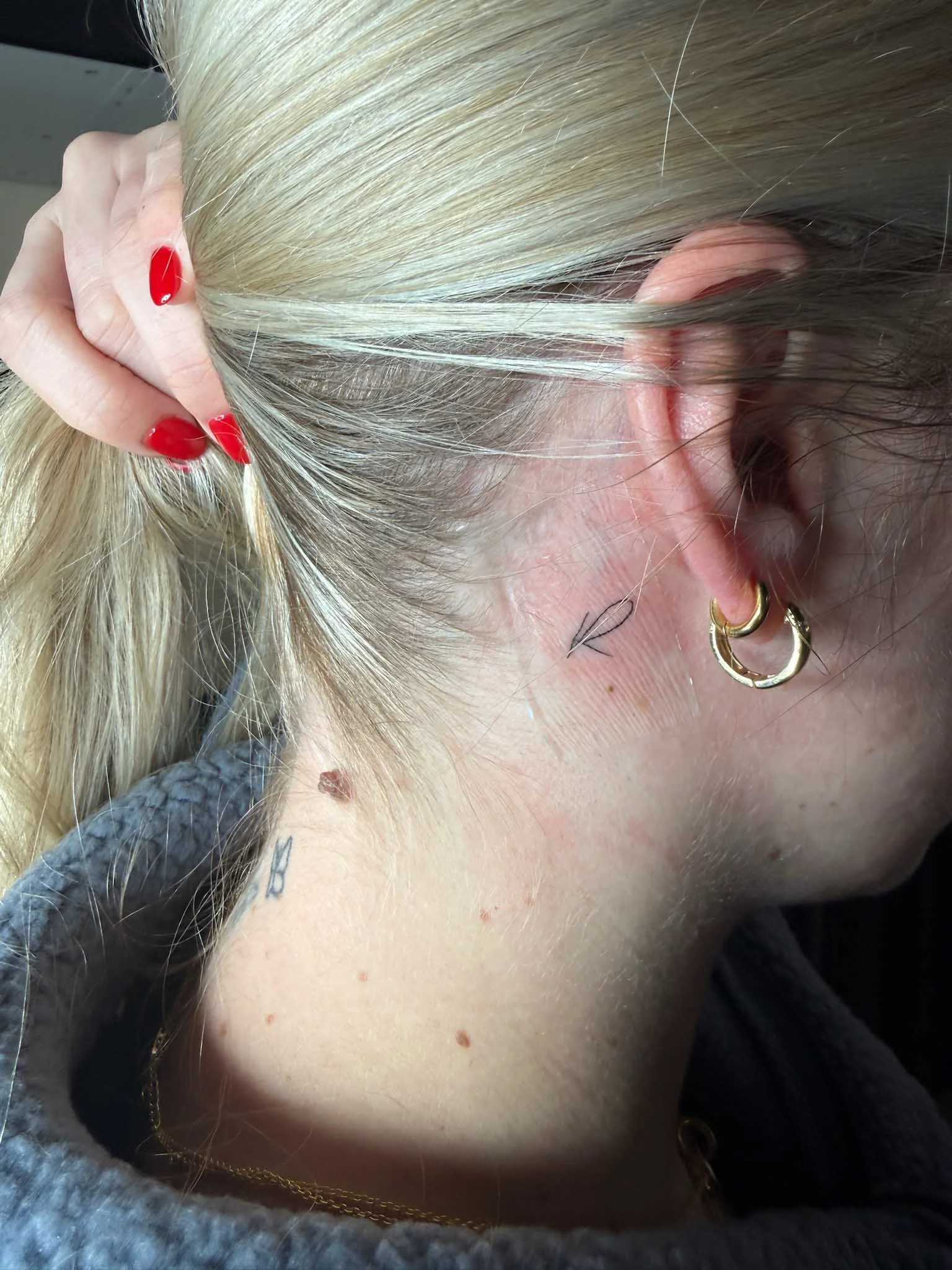 Close-up of a woman's head turned to the side, showing her blonde hair, ear with a gold hoop earring, holding her hair with her left hand with red nail polish, and a tattoo on her neck.