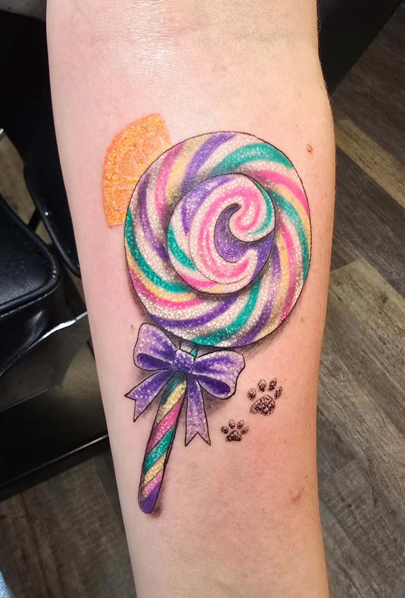 Colorful tattoo of a swirling lollipop with purple, pink, green, yellow, and white stripes, decorated with a purple bow at the stick and small paw prints nearby.
