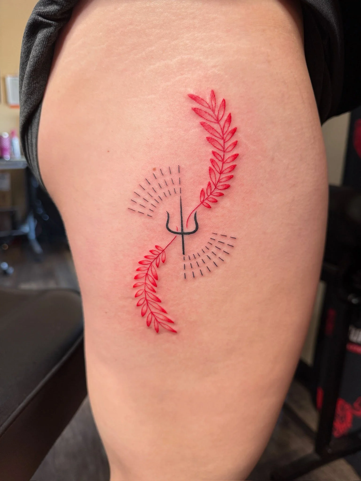 A tattoo on a person's thigh featuring a black trident with red and black botanical elements and dotted semicircular designs.