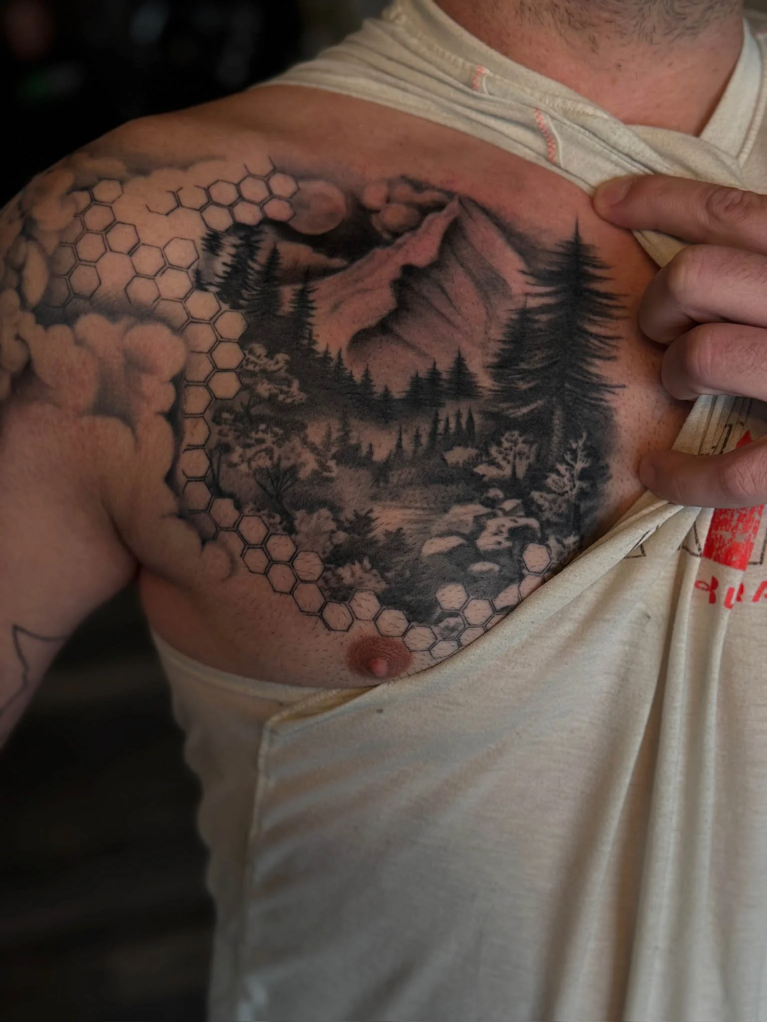 Tattoo of a mountain landscape with pine trees and clouds on a person's shoulder and chest, partially covered by a sleeveless shirt.