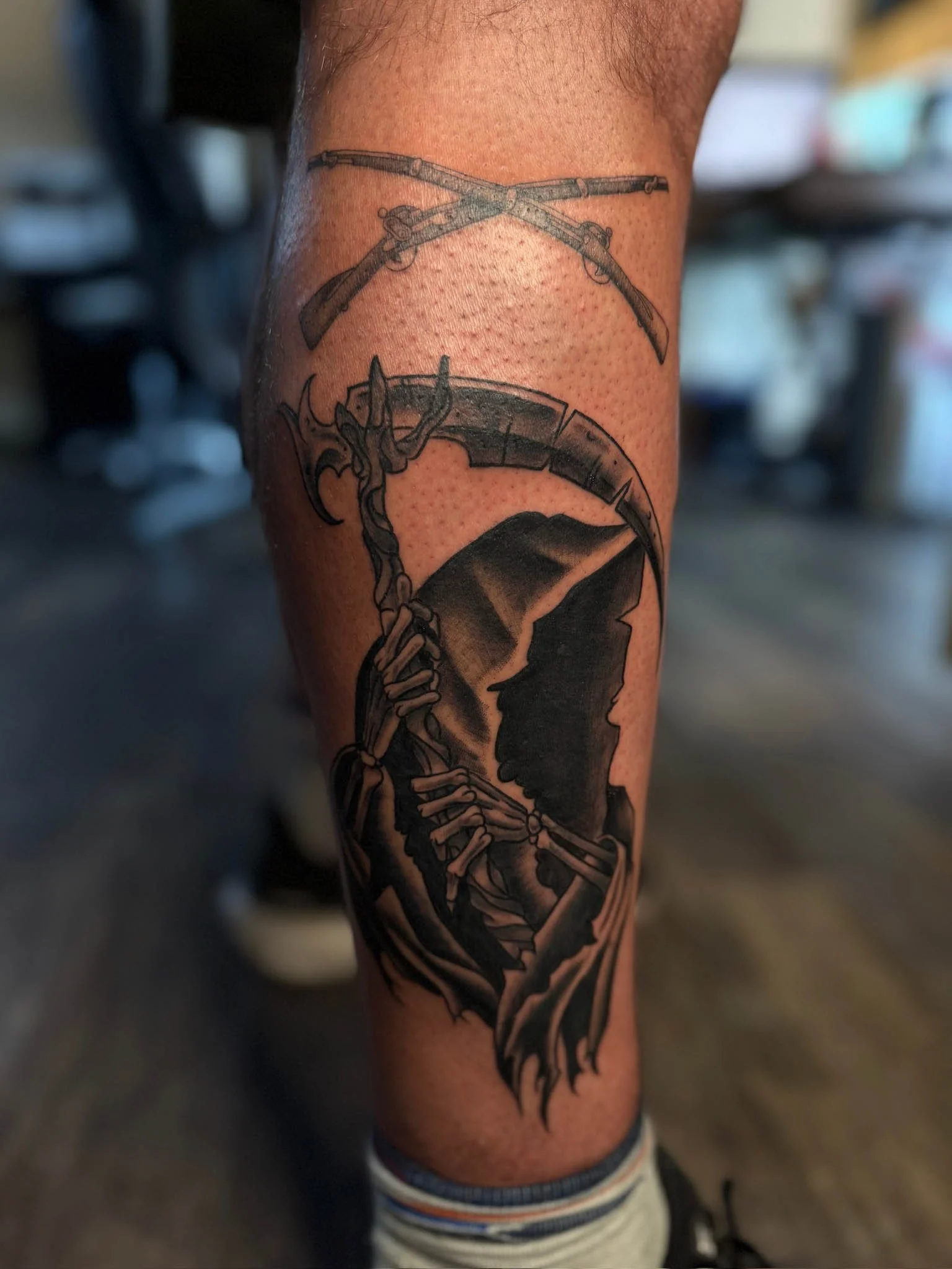 Tattoo of a Grim Reaper holding a scythe, with a silhouette of a person's face in profile, on a person's calf.