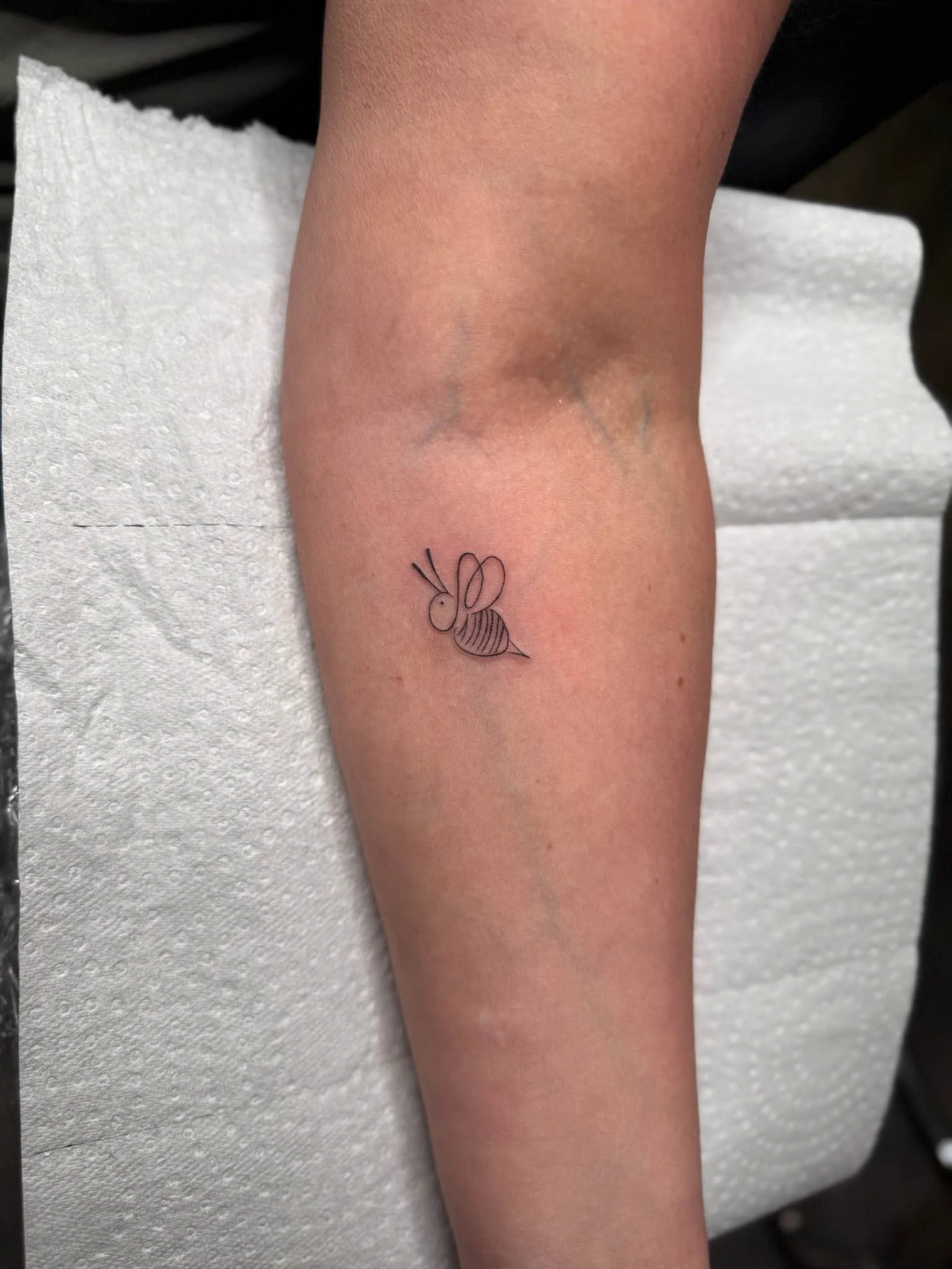 A small, simple tattoo of a bee on a person's inner forearm.