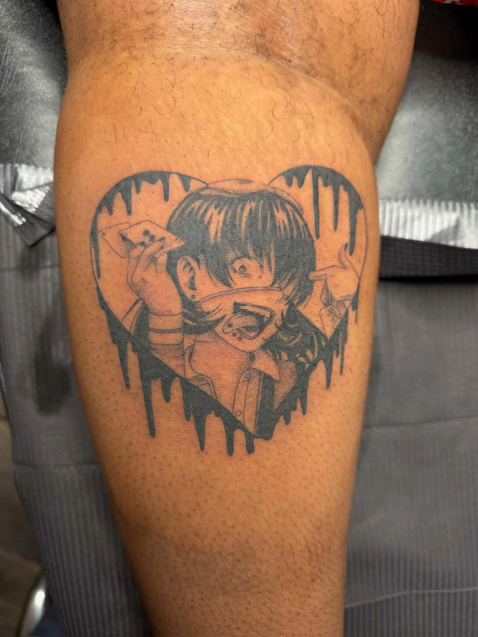Tattoo of a cartoon girl with short hair and a wide, excited expression, holding a card in one hand and making an animated gesture with the other, inside a heart-shaped outline with drippy edges.