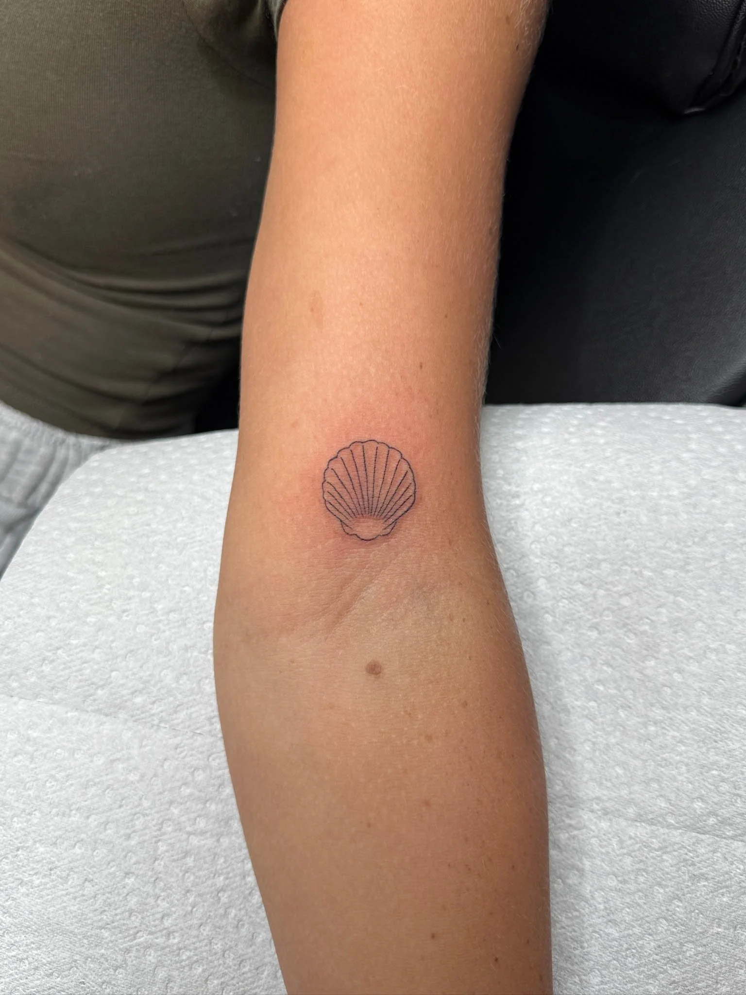 A person's inner forearm with a small shell-shaped tattoo and a tiny mole beneath it, resting on a white paper or cloth surface.