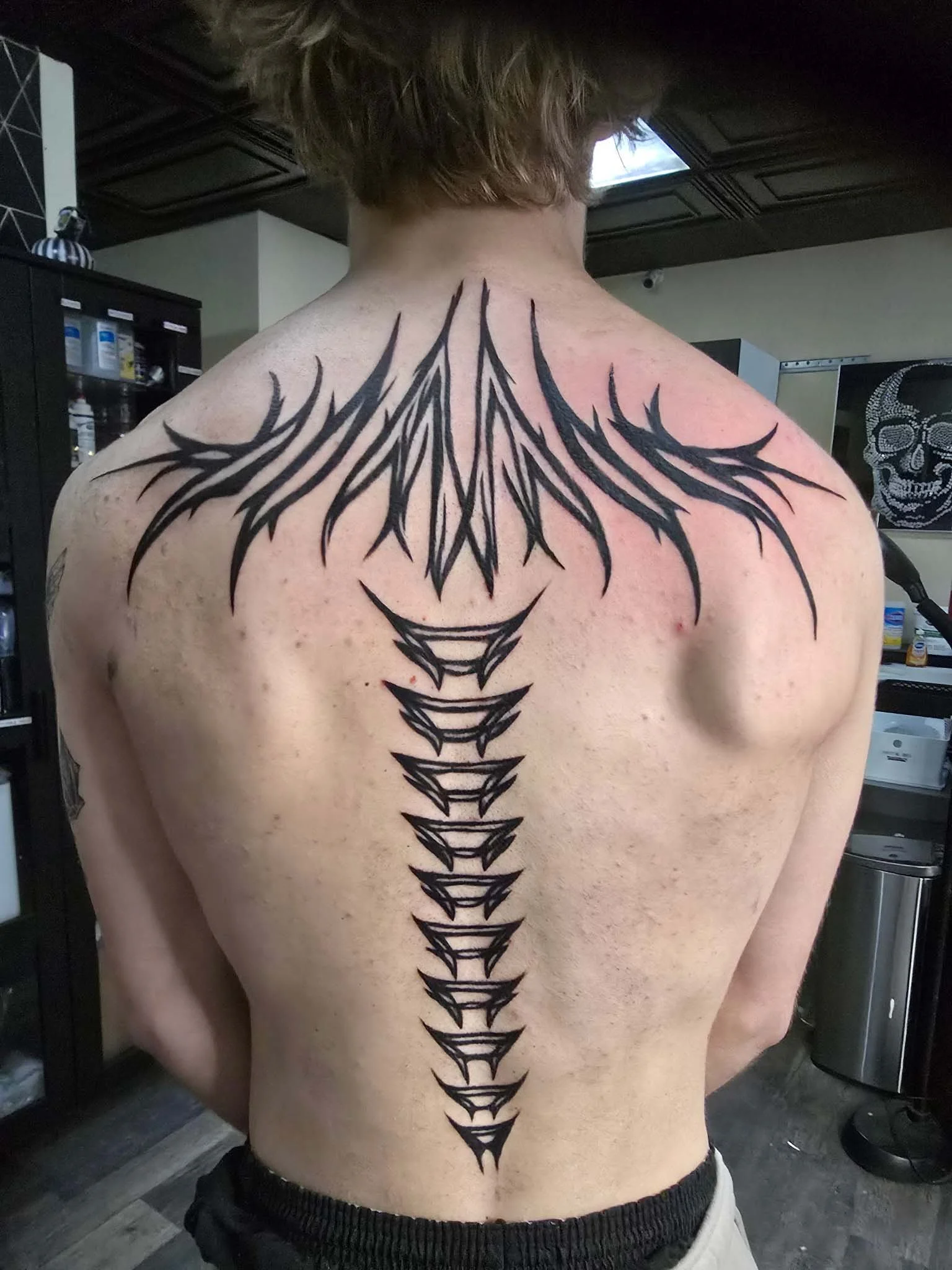 A person with a large black tribal-style tattoo running down their back, spanning from shoulders to lower back, with sharp, pointed designs and a central spine-like pattern.