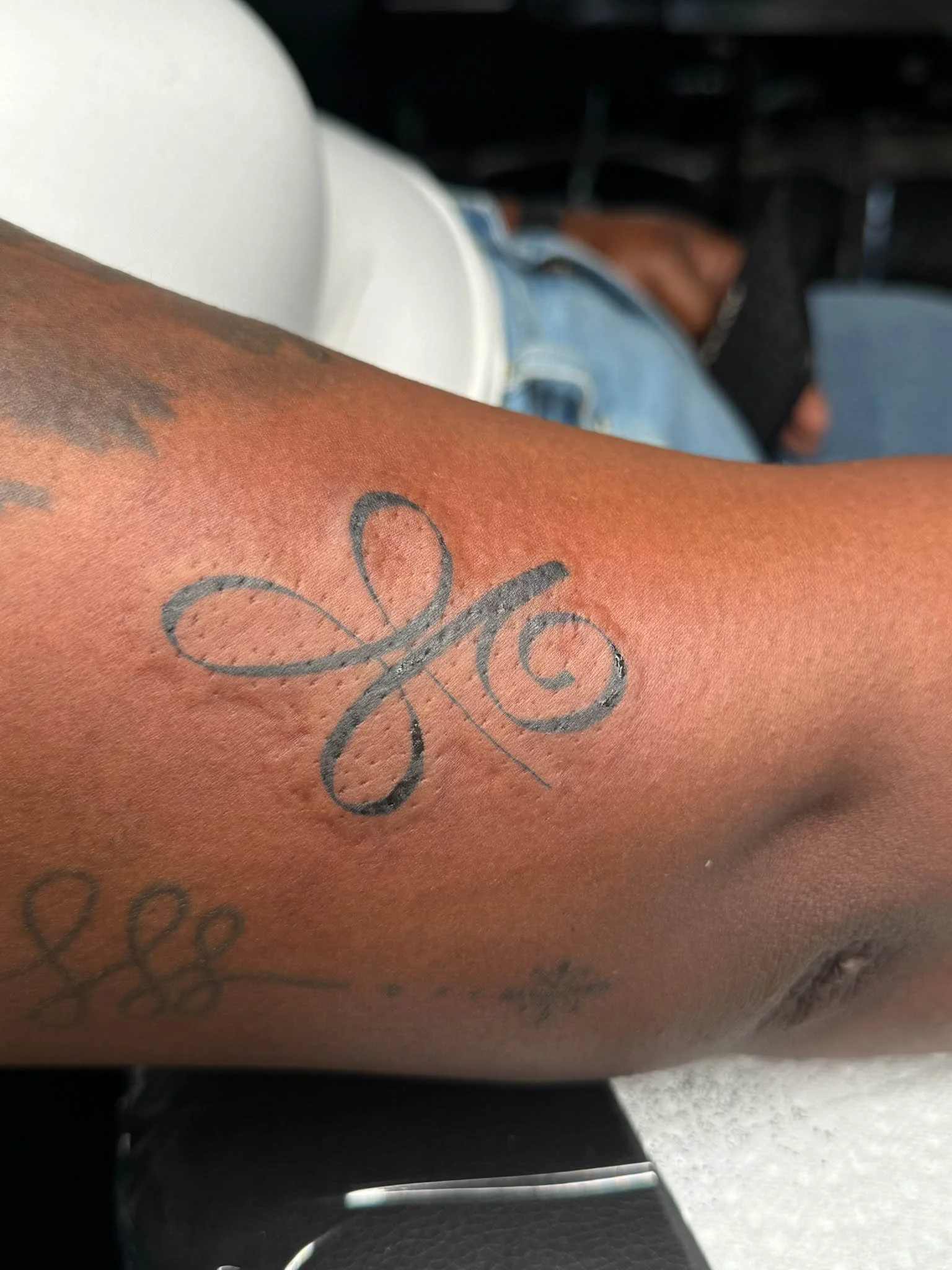 Close-up of a person's arm with tattoos, including a black outline of a butterfly and other designs, with a person lying on their side in the background.