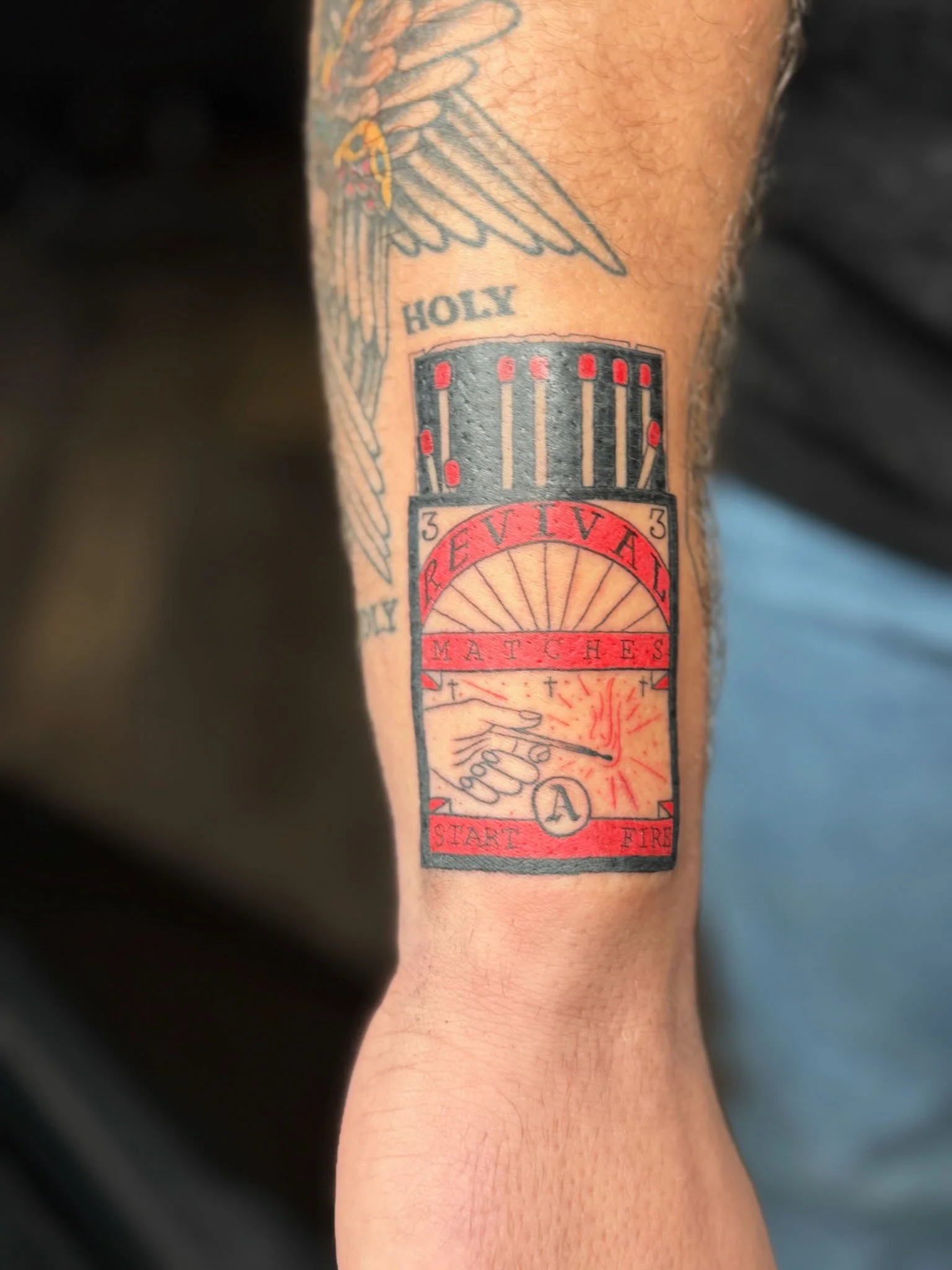 Tattoo of a matchstick with the words 'REVEV' and 'MATCHES,' a fire starting at the match's tip, and the words 'START FIRE' and 'HOLY' on the arm.
